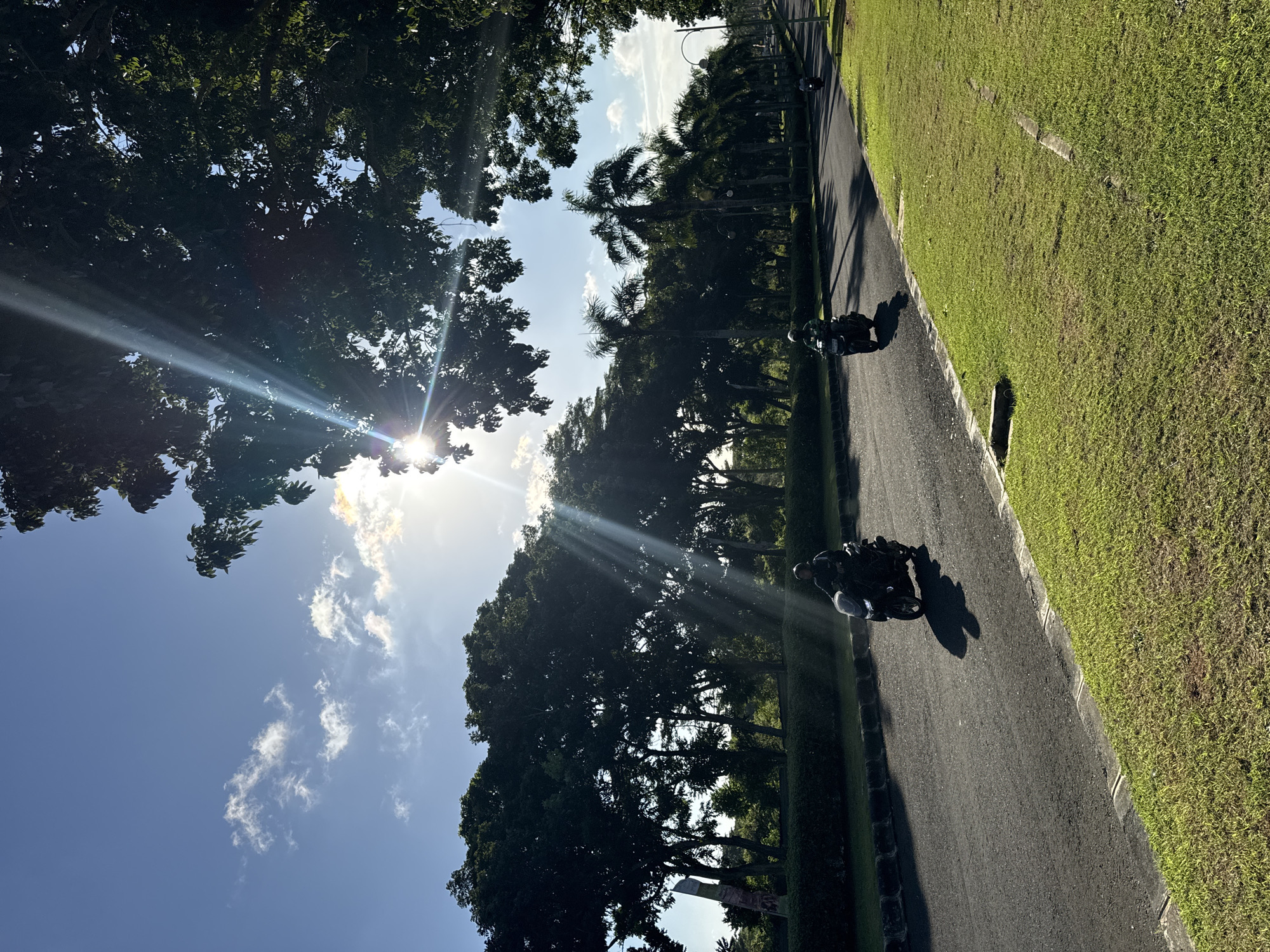 A sunny day scene in Kuta Selatan, Indonesia shows a road lined with trees and grass. Two people on motorcycles are riding on the road. The sun shines brightly through the trees, casting shadows. The sky is blue with some clouds.