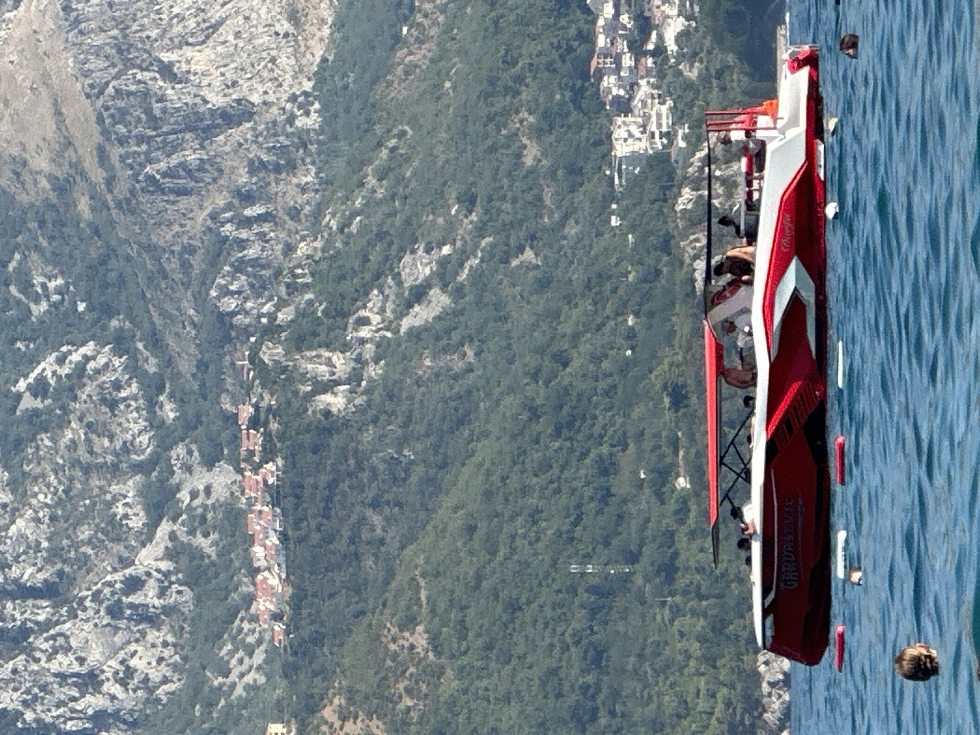 The image depicts a bright, sunny day in Boreti, Montenegro. In the foreground, the turquoise waters of the Adriatic Sea are visible, with several people swimming and their heads just above the surface. A striking red and white speedboat, identified by the name "GARDASEVIC" on its side, cuts through the water, carrying passengers. In the background, lush green hills rise steeply, dotted with trees. Clinging to the hillsides are clusters of houses with red-tiled roofs, characteristic of Mediterranean architecture. Further up, the rugged, rocky slopes of the mountains dominate the skyline. The scene conveys a sense of leisure and a typical summer day by the sea in a picturesque coastal town.