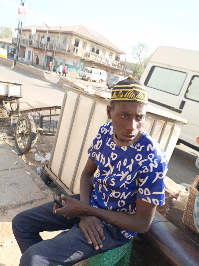 The image depicts a street scene in Gawuna, Nigeria, under bright daylight. In the foreground, an adult male with dark skin is seated on a green plastic crate. He is wearing a blue short-sleeved t-shirt with scattered white letter patterns, dark trousers, and a black kufi-style cap with yellow geometric designs. A light-colored beaded necklace is visible around his neck, and a thin green bracelet is on his left wrist. He holds a black smartphone in his right hand, with his left hand resting on his lap. His gaze is directed towards the viewer's left. Behind the man, a structure composed of light-colored panels and a metal frame is partially visible. To the man's left, a two-wheeled pushcart with a wooden-spoked wheel rests on the ground. The background features a paved street ascending gently, lined by a multi-story building with balconies and other structures. Multiple vehicles, including a white van prominent in the mid-ground and other vehicles further down the road, are present. Several indistinct individuals are visible walking in the distant background.