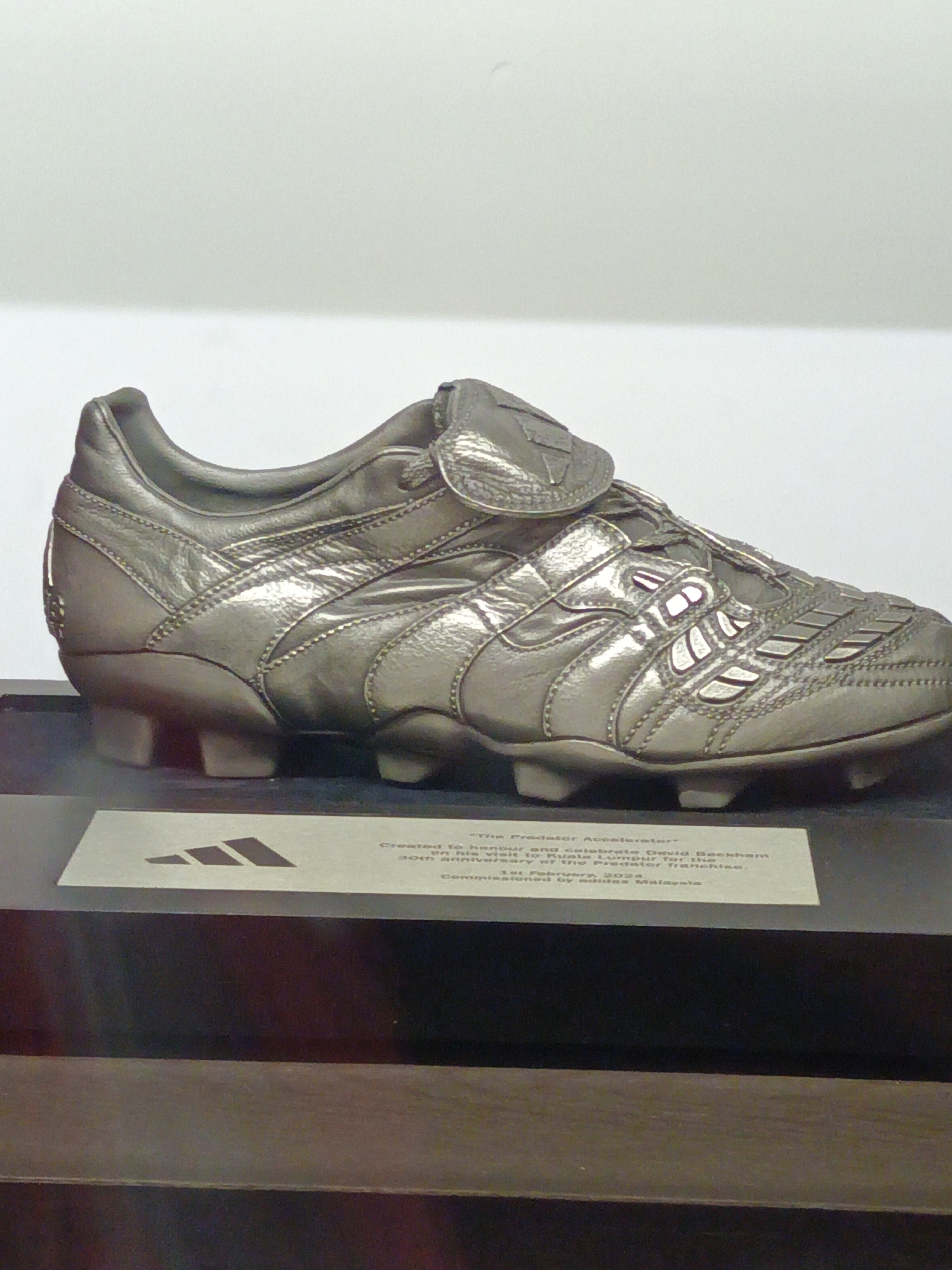 The image shows a silver metallic sculpture of an Adidas Predator Accelerator football boot, displayed on a black pedestal. A plaque is placed in front of the boot, bearing the Adidas logo and text that reads "The Predator Accelerator" created to honor and celebrate David Beckham's visit to Kuala Lumpur for the 30th anniversary of the Predator franchise, commissioned by Adidas Malaysia, dated 1st February 2024. The background is a plain, light-colored wall. The scene is indoors, likely in a museum or exhibition space in Kuala Lumpur, Malaysia, celebrating a significant milestone for the Adidas Predator football boot.