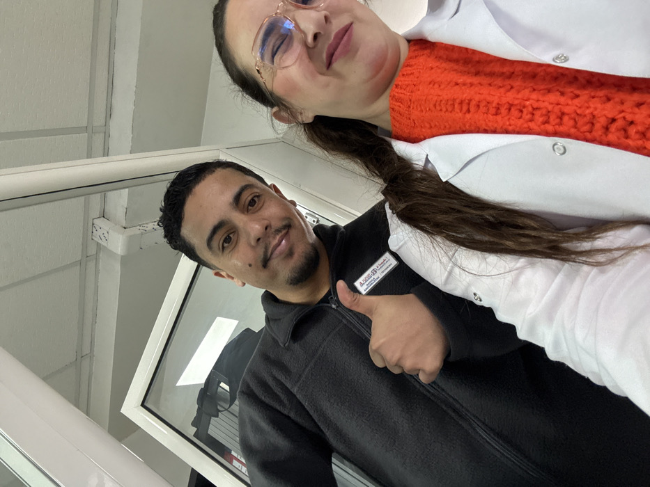 The image displays two individuals, one male and one female, posing in an indoor commercial or professional setting within Meknes, Morocco. The male, positioned to the left, has dark hair, a mustache, and a short beard. He is wearing a dark, zippered fleece jacket with a white identification badge on his right chest, which reads "A. Hajjaj & F." and additional Arabic text. He presents a faint smile and a "thumbs up" gesture with his right hand. The female, positioned to the right, has dark hair tied back and is wearing light-framed eyeglasses. She is smiling and dressed in a bright orange, cable-knit sweater underneath a white lab coat or professional jacket. The background consists of a white ceiling with exposed conduits and a white-framed door or window to the left. The environment is brightly illuminated by artificial lighting.