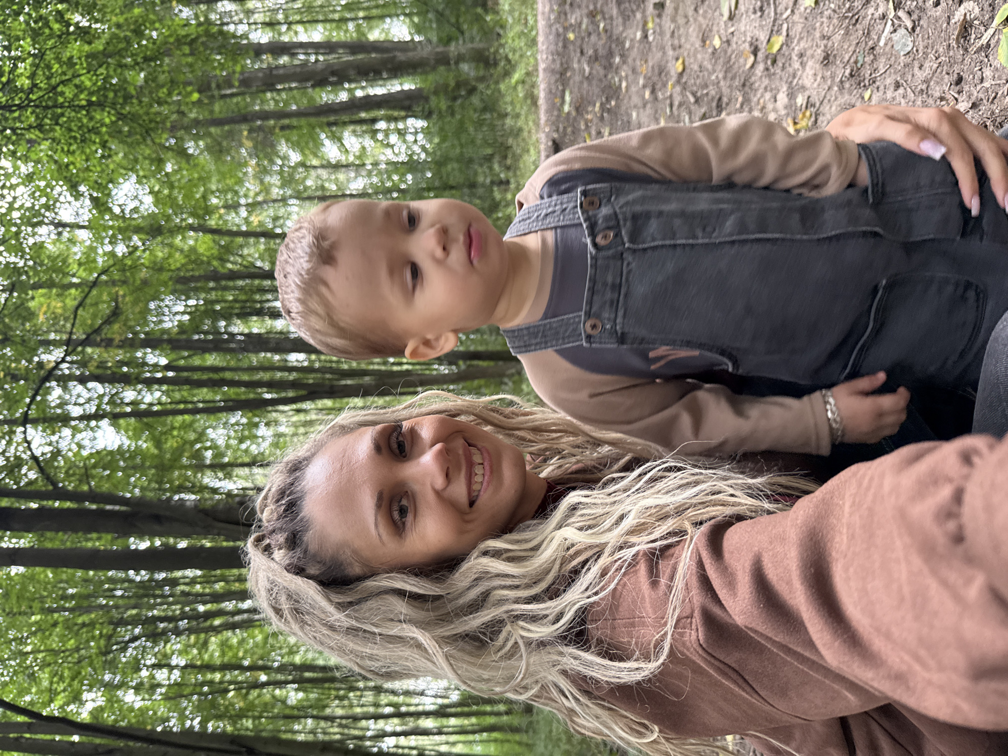 A woman with blonde, curly hair pulled into a ponytail is taking a selfie with a young child in a forest. The woman is wearing a brown jacket and smiling at the camera. The child is wearing a tan long-sleeved shirt and grey overalls, and is looking away from the camera. The background consists of tall trees with green foliage, suggesting it is a wooded area. The ground is covered with dirt and fallen leaves. The location is described as the city of Dumino, Russia.