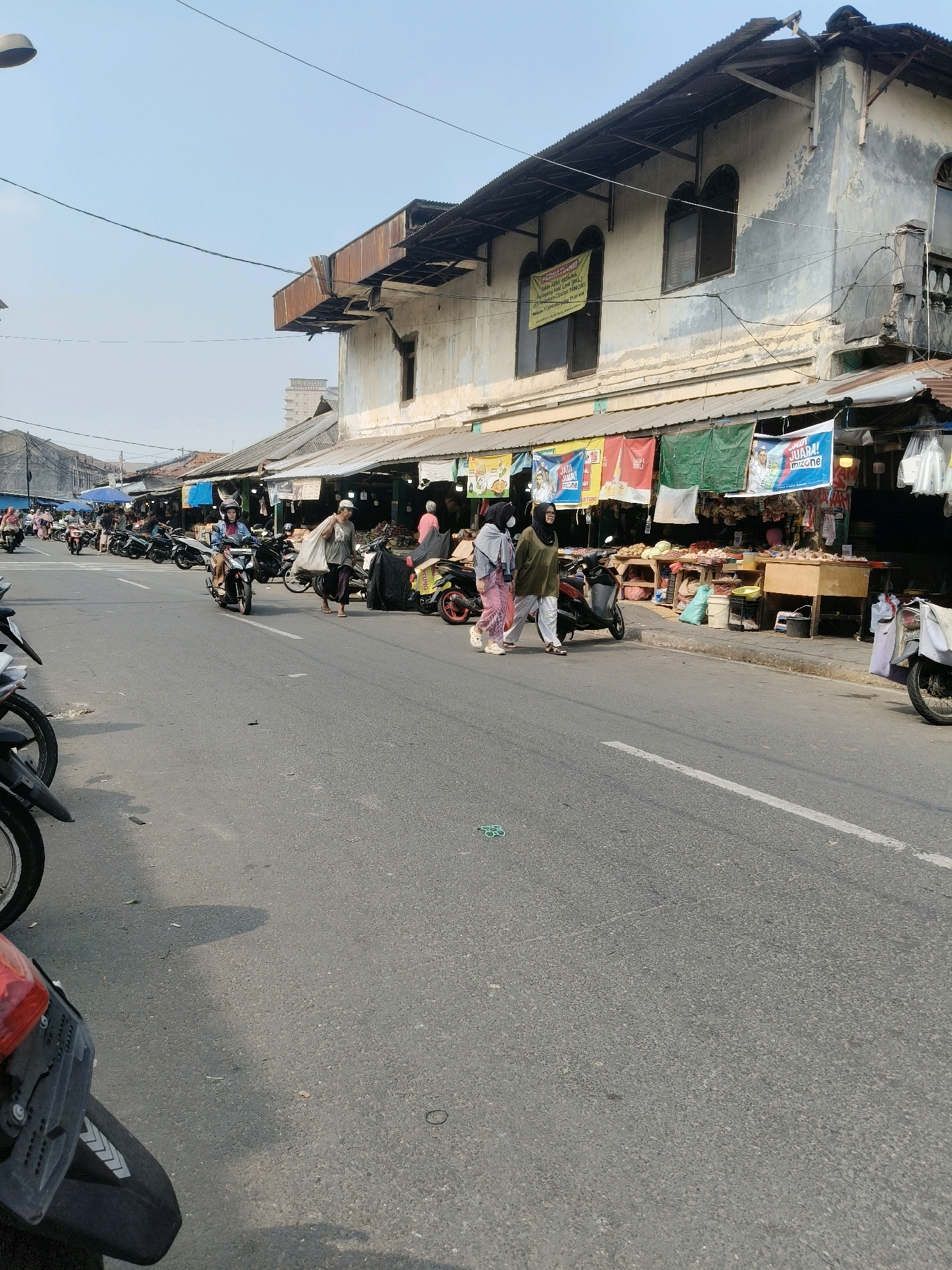 The image depicts a street scene in South Tangerang, Indonesia, likely a market area. The foreground is dominated by the asphalt road with several parked motorcycles and a portion of a motorcycle in the lower left corner. In the middle ground, a street market is set up along the side of the road, with stalls displaying various goods, possibly food items, under awnings. Several people are walking along the market area and crossing the street. One person is walking across the street carrying a large bag over their shoulder, while two women in headscarves are walking together. Several motorcycles are moving or parked, indicating active commerce and transportation. The background shows a long building with an aged facade, featuring multiple windows and a weathered appearance, suggesting it is an older structure. The sky is clear and bright, indicating daytime. The overall scene conveys a busy, everyday market environment.