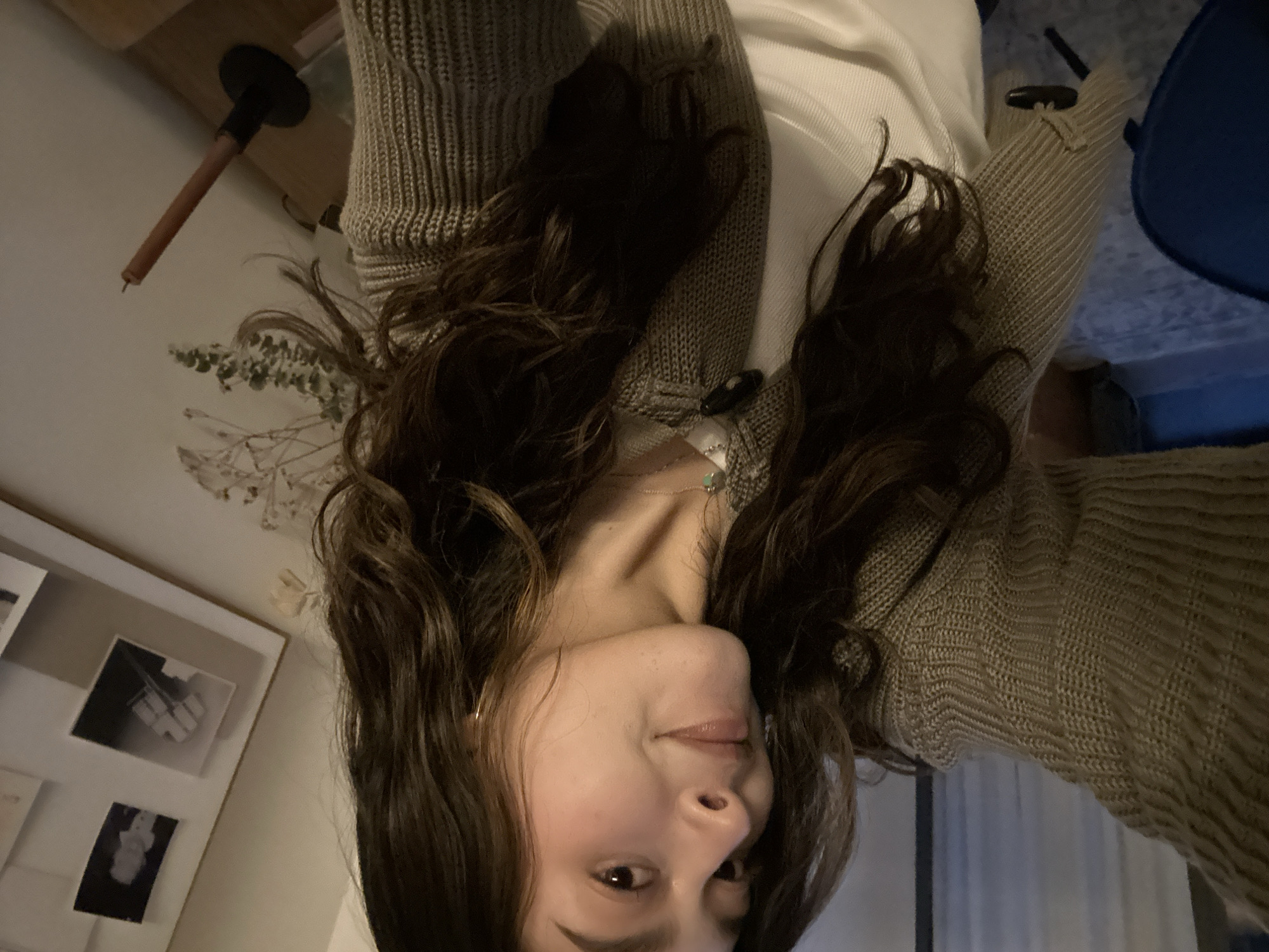 A rotated indoor shot depicts a woman with long, dark brown, wavy hair. She is wearing a light-colored knit sweater over a white shirt. The woman's face is visible, and she appears to be looking at the camera. In the background, a picture frame with multiple black and white photos is hung on the wall. A wooden table with a black lamp is seen in the upper left corner. The location cannot be determined from the image.