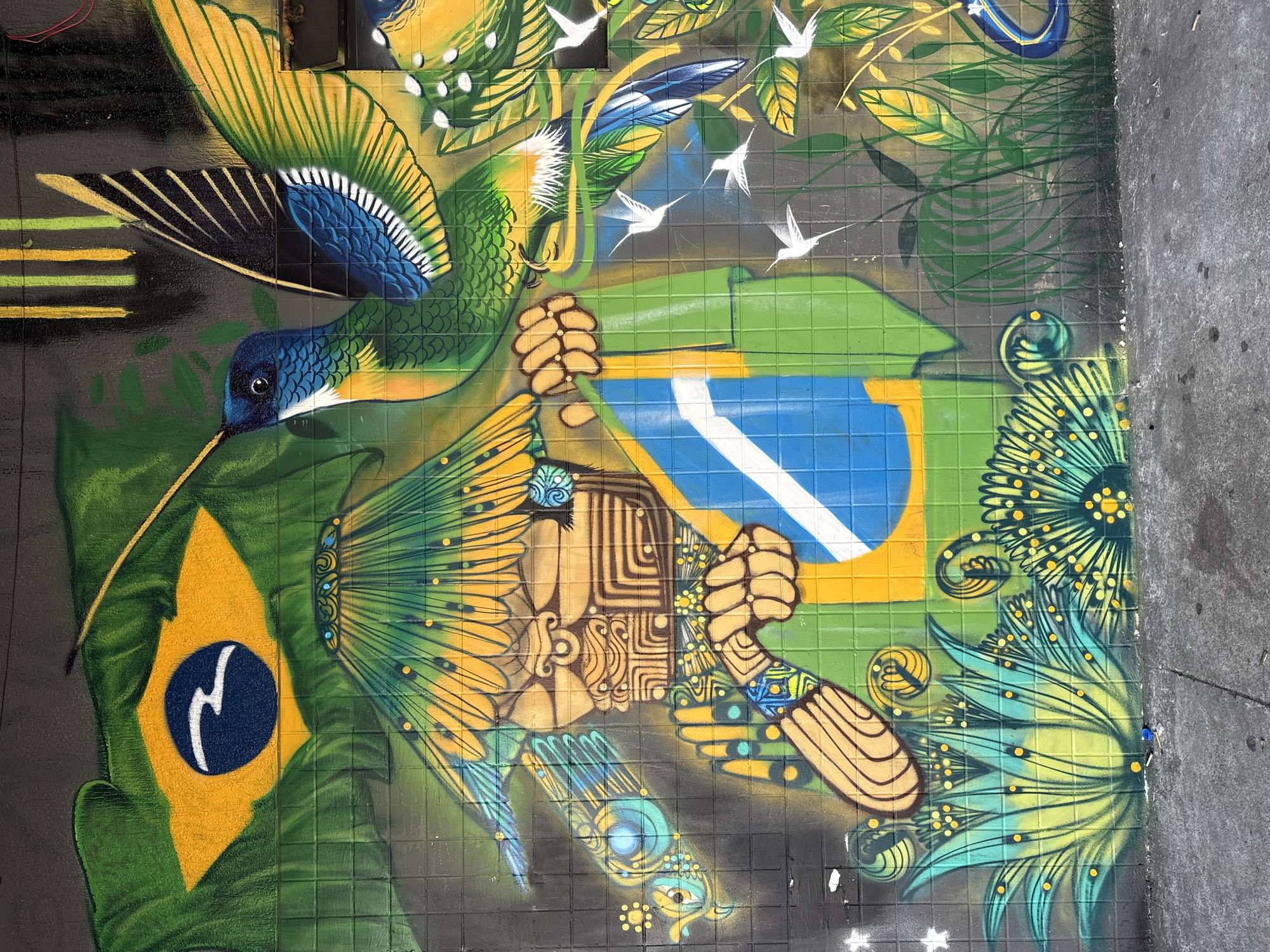 The image displays a vibrant graffiti mural on a tiled wall in São Paulo, Brazil. The mural features a stylized depiction of the Brazilian flag, with its green background, yellow rhombus, and blue circle containing white stars and a white band. A hummingbird with blue, green, and yellow plumage is prominently featured, appearing to fly towards the flag. To the left of the flag, a figure with indigenous-inspired headdress and facial markings is rendered in earthy tones. The figure's hands hold the Brazilian flag. The mural also includes lush green foliage, depicted with yellow and blue accents, and several smaller white birds, possibly also hummingbirds, scattered throughout the scene. The overall composition suggests a celebration of Brazilian culture and nature.