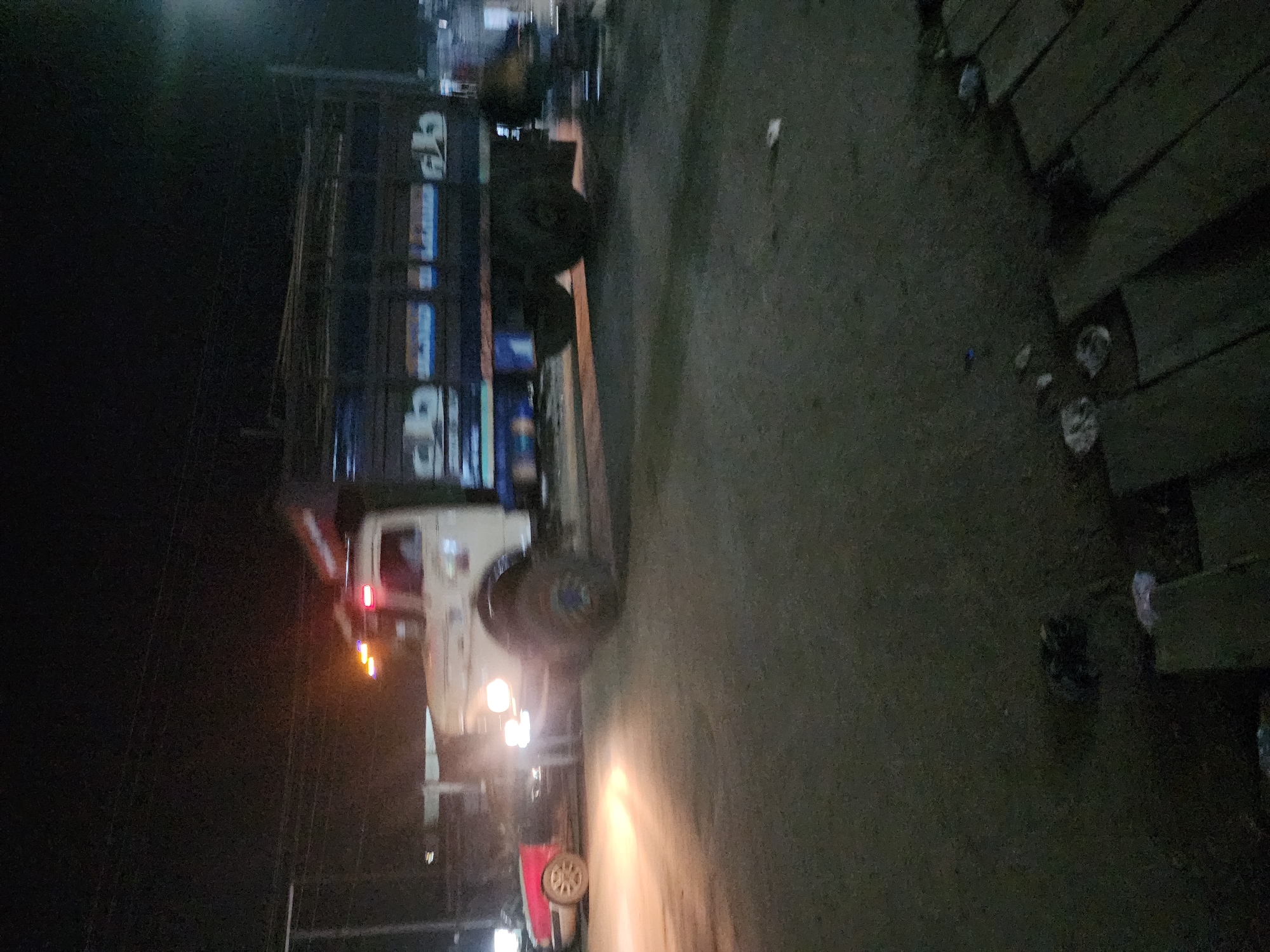 A blurry image depicts a vehicle with a trailer on a road, possibly at night in Obiaruku, Nigeria. The trailer has signage in an unknown language. The environment is dark and indistinct, with limited visibility. The image appears to be taken from a low angle.