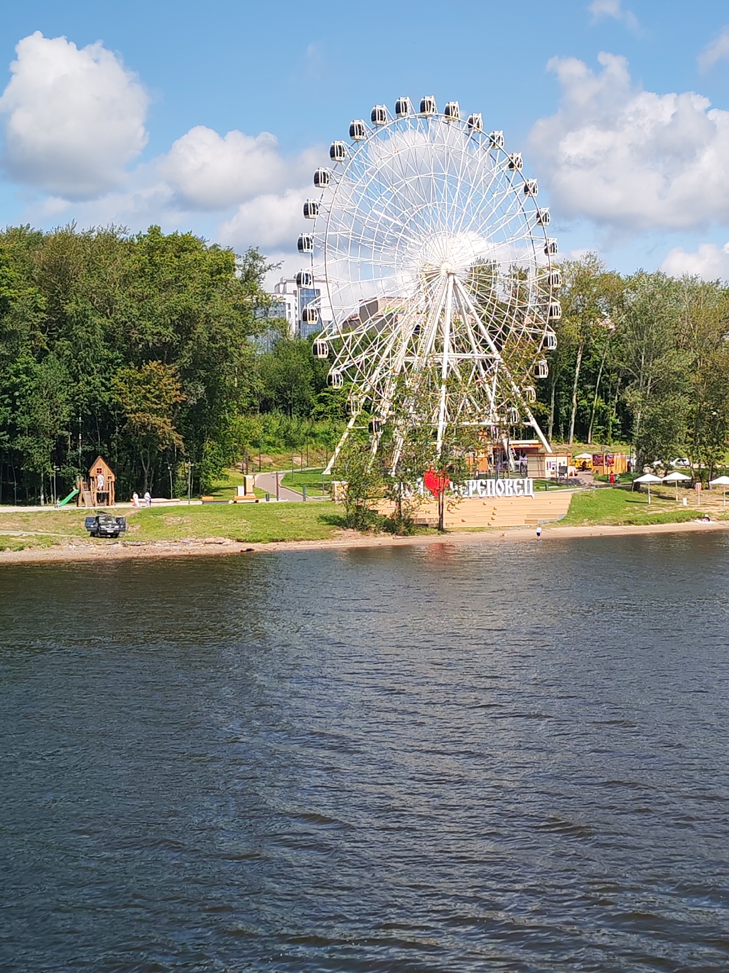 The image depicts a scenic view of the city of Cherepovets, Russia, with a prominent Ferris wheel as the central subject. The Ferris wheel, a large white structure with numerous enclosed cabins, dominates the background, set against a clear blue sky with scattered white clouds. The Ferris wheel appears to be located within an amusement or recreational area, as indicated by the presence of trees, grass, and a body of water in the foreground.
In the mid-ground, a sandy shoreline is visible, with people relaxing on the beach and children playing near a wooden playground structure. A dark-colored vehicle is parked on the sand near the water's edge. Several white umbrellas are set up on the beach, providing shade. Behind the beach, there is a grassy area with walking paths and a sign with Cyrillic lettering that appears to spell out "ЧЕРЕПОВЕЦ" (Cherepovets).
The foreground is occupied by a dark, rippling body of water, likely a river or lake, reflecting the blue sky and the surrounding greenery. The overall impression is one of a lively, outdoor recreational space on a sunny day.