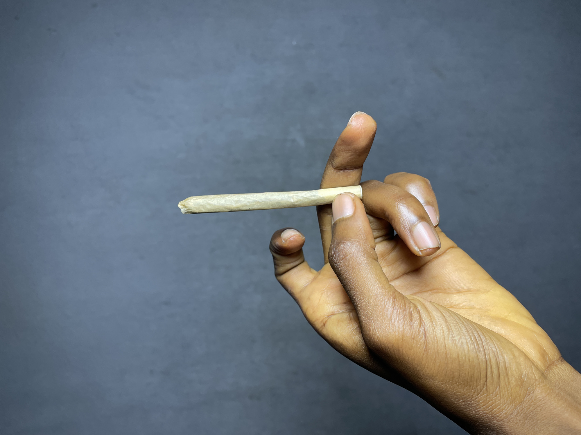 A close-up, waist-up, medium shot shows a person's dark-skinned hand holding a rolled cigarette between their thumb and index finger. The cigarette is pale yellow and slightly unrolled at the tip. The background is a dark, out-of-focus gray.