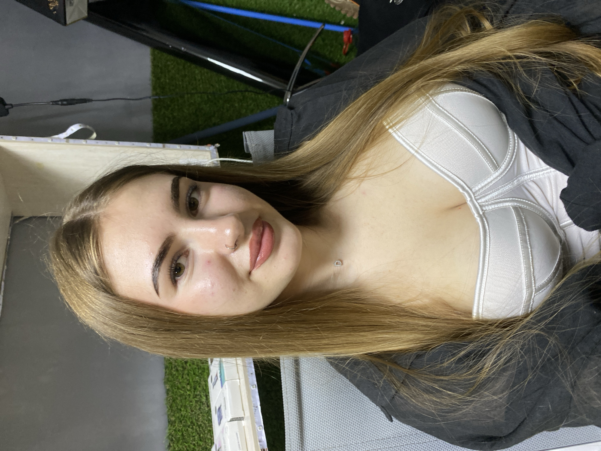 A young woman with long, blonde hair is looking at the camera. She is wearing a white, structured top and a dark jacket. She has a nose ring and a small necklace with the letter "D" on it. The background appears to be indoors, with a shelf holding several boxes and artificial green turf visible. The scene is a close-up shot of the woman's upper body. There is no identifiable information regarding the specific location within the city of Dyakovo, Russia.