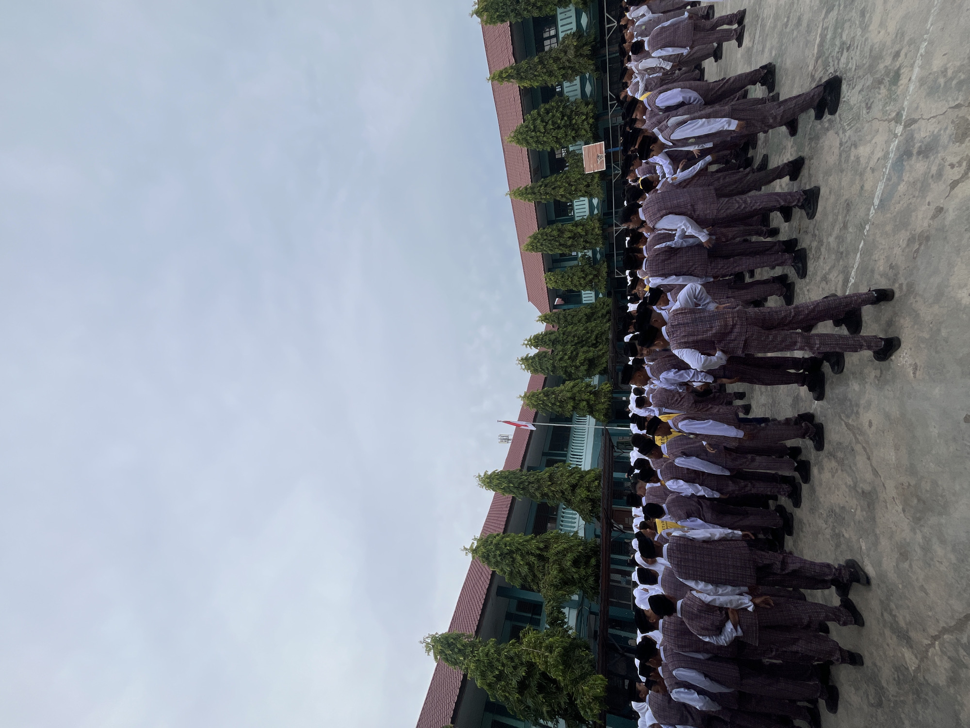 The image shows a large group of students, likely in uniform, standing in an outdoor area in front of a two-story school building. The students appear to be young males, and many are wearing plaid vests over white shirts, with dark pants and black caps. Some students are wearing headscarves, suggesting there might be female students present as well, though the majority visible are male. They are standing on a paved surface, possibly a schoolyard or plaza. The building behind them has a red tiled roof and a light green facade with balconies and windows. Several tall, slender trees line the front of the building. A red and white flag, likely the Indonesian flag, is visible flying from a flagpole. The sky is overcast. This scene is consistent with a school assembly or outdoor class activity. Based on the presence of the Indonesian flag and the architectural style of the building, it is plausible this scene is located in Lhokseumawe, Indonesia.