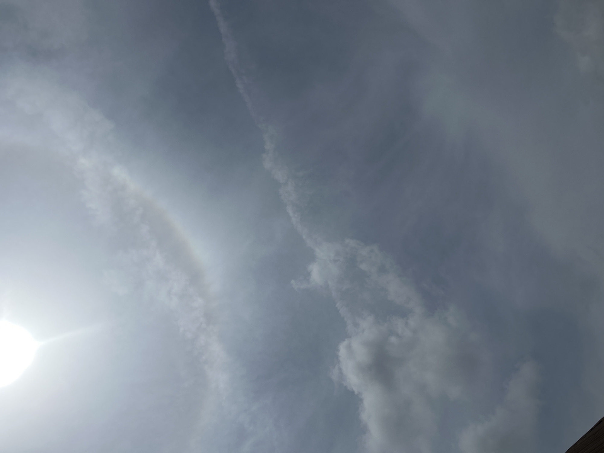The image shows a sky with clouds and the sun partially visible. There is a sun dog, which is a circular halo around the sun caused by ice crystals in the atmosphere. The sky appears to be overcast, with patches of both light and dark clouds. The sun is bright and shining, creating a lens flare effect in the upper left corner of the image. There is a faint rainbow visible in the lower right portion of the image, indicating atmospheric conditions conducive to its formation. The foreground of the image shows a portion of a brown roof, suggesting the photograph was taken from a vantage point on a building. The overall scene is natural and depicts a weather phenomenon. The image was taken in Ogbomosho, Nigeria.