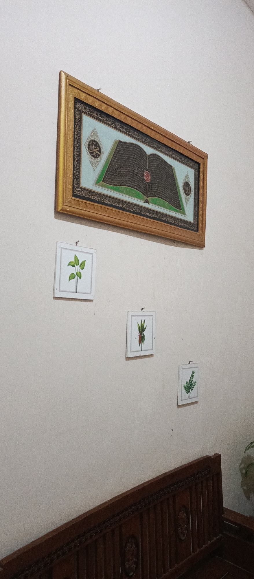 A rectangular, framed artwork hangs on a white wall. The artwork depicts an open book with dark text on a light blue background, framed by a gold border. Below this main artwork are three smaller, square framed artworks, also hanging on the wall. The first, to the left, shows a green plant with multiple leaves. The second, to the right and slightly lower, features a red object with green leaves. The third, further to the right and lower still, displays a green leafy plant. In the foreground, a dark wooden bench with ornate carvings is visible. The scene appears to be indoors, likely in a home or a communal space.