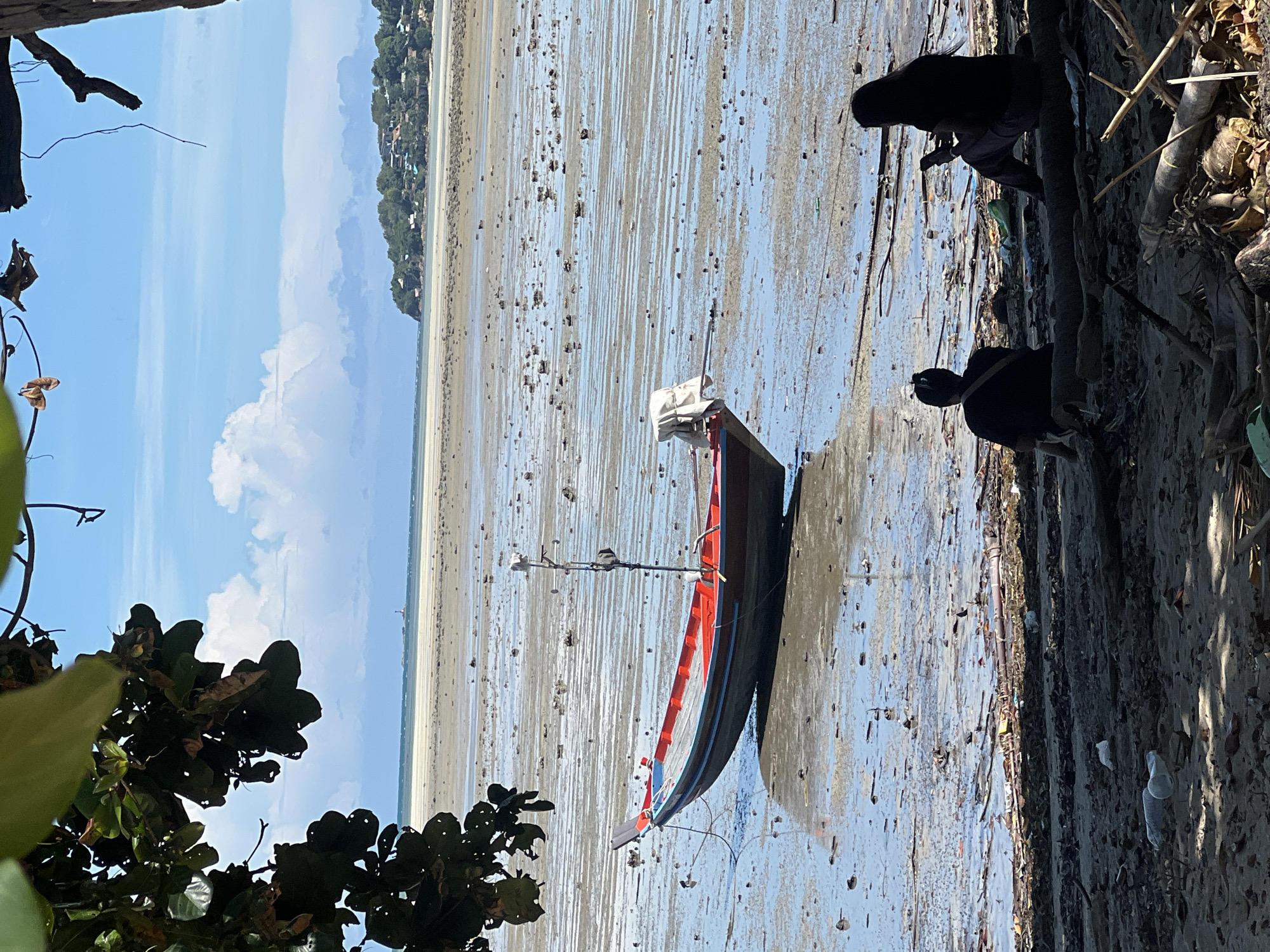 The image depicts a low tide scene on the coast of Koh Phangan, Thailand. A red and blue fishing boat is grounded on the wet sand, its hull reflecting the sky. The vast expanse of the receding tide reveals a muddy seabed littered with debris and small rocks. In the foreground, two individuals are seated on a log, facing the water. One person, in silhouette, is looking down at a phone, while the other person, also in silhouette, is sitting with their back to the viewer. Behind them, the shoreline is covered in sand, driftwood, and various forms of litter, including plastic bottles. In the distance, a treed hillside with buildings stretches along the coast, and the ocean meets a hazy horizon under a partly cloudy sky.