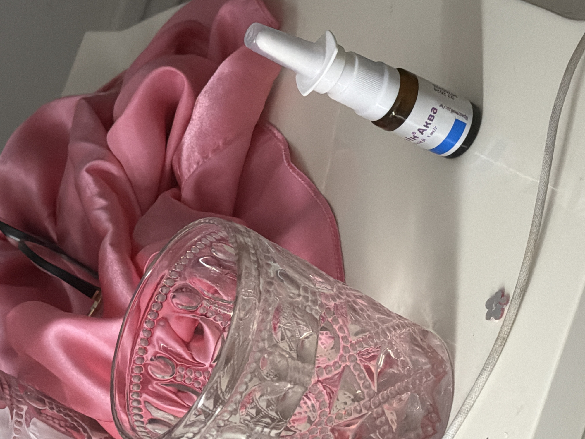 This is an indoor photograph featuring a collection of personal items arranged on a white surface, possibly a shelf or table. The background is dominated by a soft, draped pink satin fabric, which appears to be a scarf or piece of clothing. To the left, a clear, ornate glass sits partially filled with liquid. In the foreground, to the right of the glass, a small brown glass bottle with a white spray nozzle is positioned. The bottle has a label with Cyrillic text and a blue stripe. A thin, light-colored cord, possibly from a charger or accessory, runs diagonally across the lower part of the frame, with a small, gray, flower-shaped embellishment near the glass. A pair of eyeglasses with dark frames is partially visible within the folds of the pink fabric. The lighting suggests an indoor setting, possibly a bedroom or vanity area. There is no information available to indicate the location in the city of Bila Tserkva, Ukraine.