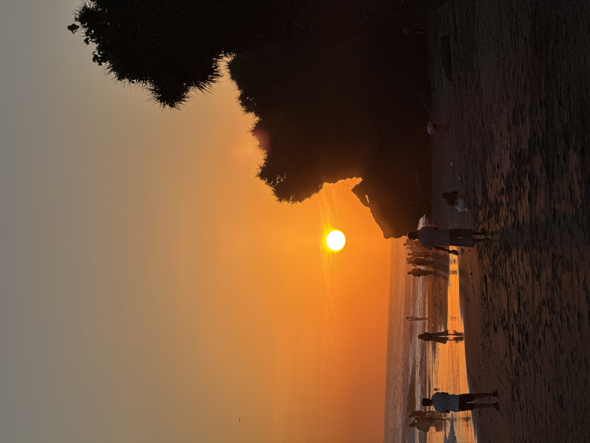 The image shows a sunset at a beach with people on the shoreline. The sun is setting over the ocean, creating an orange glow in the sky. Silhouetted figures of people walking on the beach are visible. A dark mass of vegetation overhangs part of the scene. The location is identified as Kediri, Indonesia.