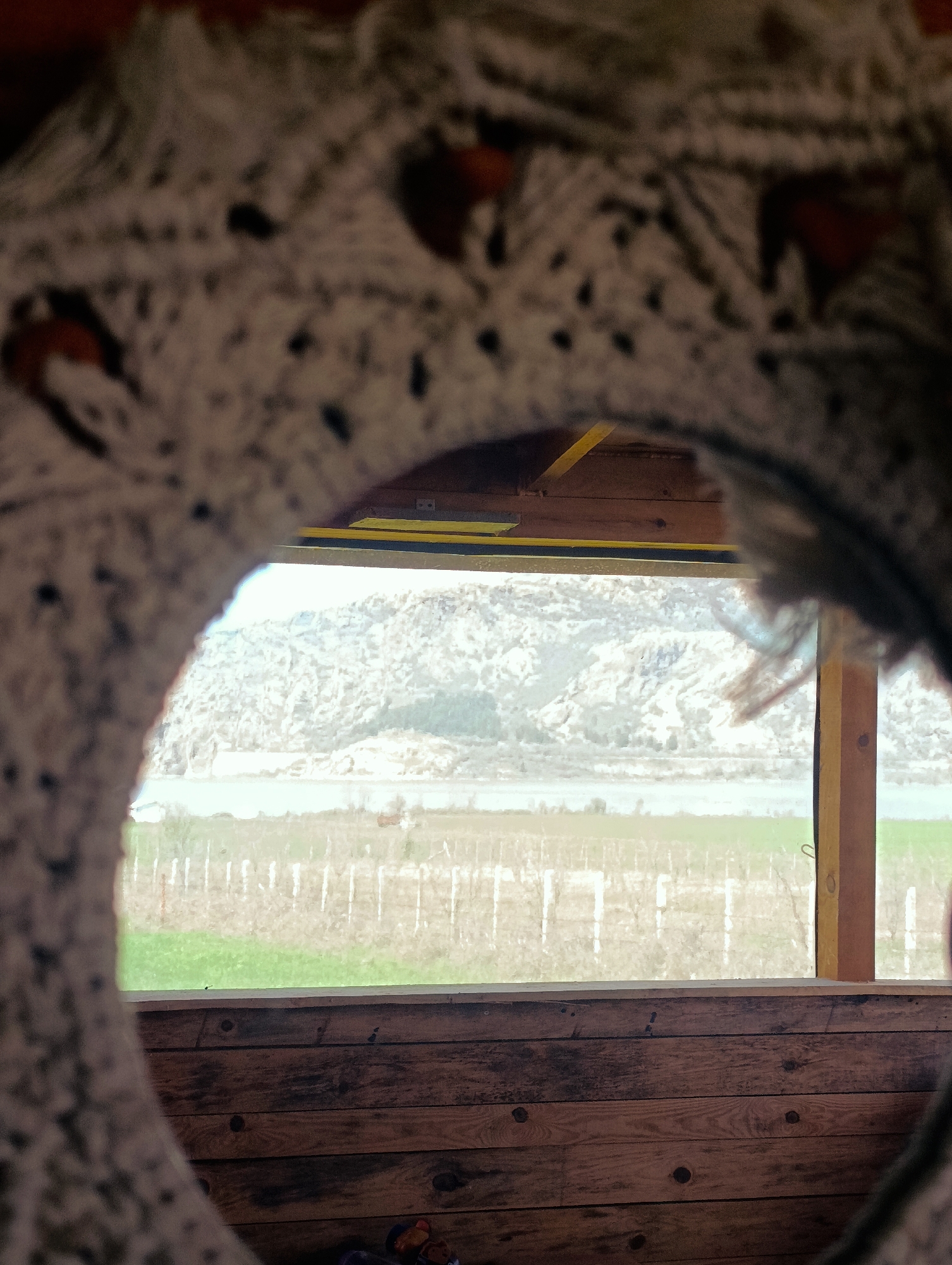 The image shows a view through a roughly circular, macrame-style frame. The view beyond is of a landscape with mountains in the background, a narrow body of water in the middle ground, and a field with a fence in the foreground. The foreground also features the wooden interior of a structure. The scene suggests an outdoor setting in the vicinity of Shiroko pole, Bulgaria, given the provided location. No people are visible in the image.