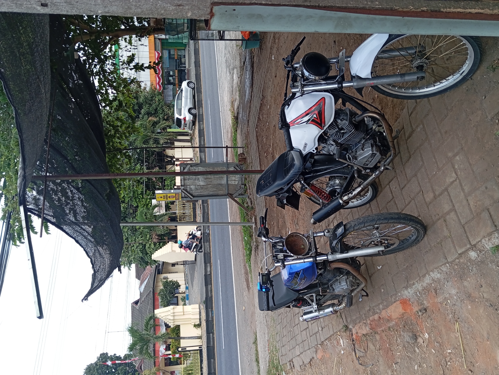 Two motorcycles are parked on a brick sidewalk next to a dirt shoulder. The motorcycle in the foreground on the left is blue with a black seat and silver exhaust pipes. The motorcycle on the right is white with red and black accents on the fuel tank and a black seat. A black sunshade is suspended overhead, supported by metal poles. In the background, a paved road runs parallel to the sidewalk, with a white car and two people on a motorcycle driving on it. Buildings line the opposite side of the road, including one with a yellow facade and white pillars. The overall environment appears to be a semi-urban area in Lemahbang, Indonesia, possibly a commercial or public service area.