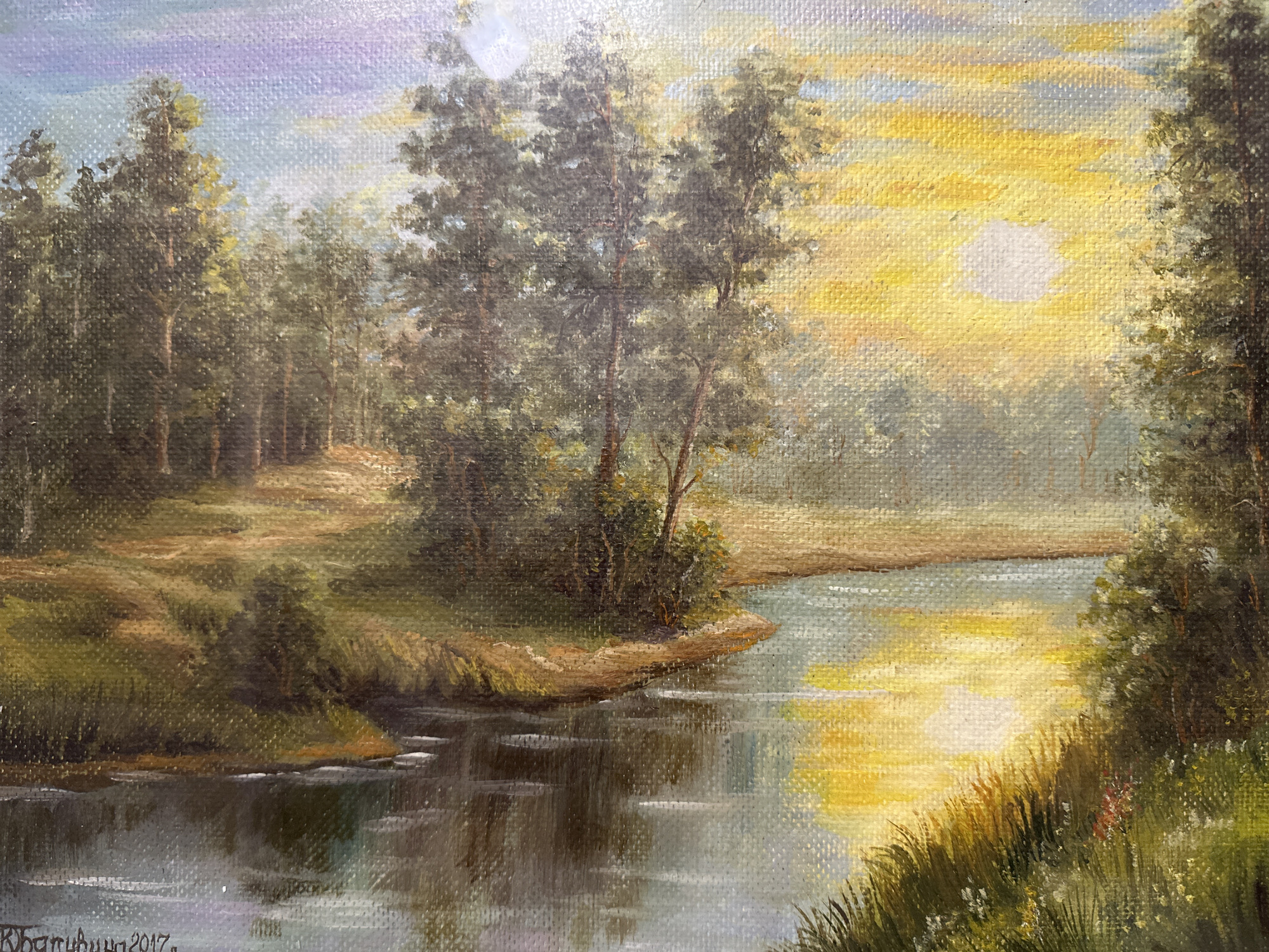 This is an oil painting on canvas depicting a serene landscape with a river flowing through a wooded area. The sun is setting or rising, casting a warm, golden light across the scene. The sky is a blend of purple, blue, and yellow hues, with clouds illuminated by the sun. Tall trees with dense foliage line the banks of the river, with some appearing to be deciduous and others evergreen. The river's surface reflects the colors of the sky and the surrounding vegetation, with gentle ripples disturbing the water. The foreground features tall grasses and some small wildflowers along the riverbank. The overall impression is one of tranquility and natural beauty. The signature at the bottom left corner reads "К. Батюшкова 2017", indicating the artist and the year of creation. There are no people visible in the painting. The painting does not contain any specific elements that identify it as being located in Podolsk, Russia.