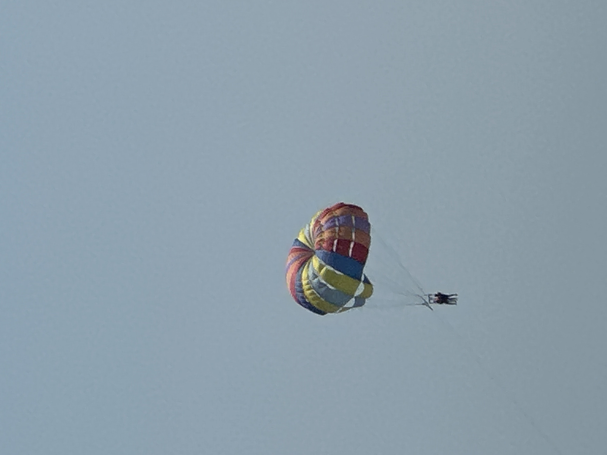 A colorful parachute with red, orange, yellow, green, and blue stripes is suspended in a light blue sky, carrying two people. The parachute is positioned in the middle right of the frame. The two people, appearing as small dark figures, are hanging beneath the parachute. The sky is clear and overcast, suggesting it is daytime. No discernible city or land features are visible, thus location in Boreti, Montenegro cannot be confirmed.