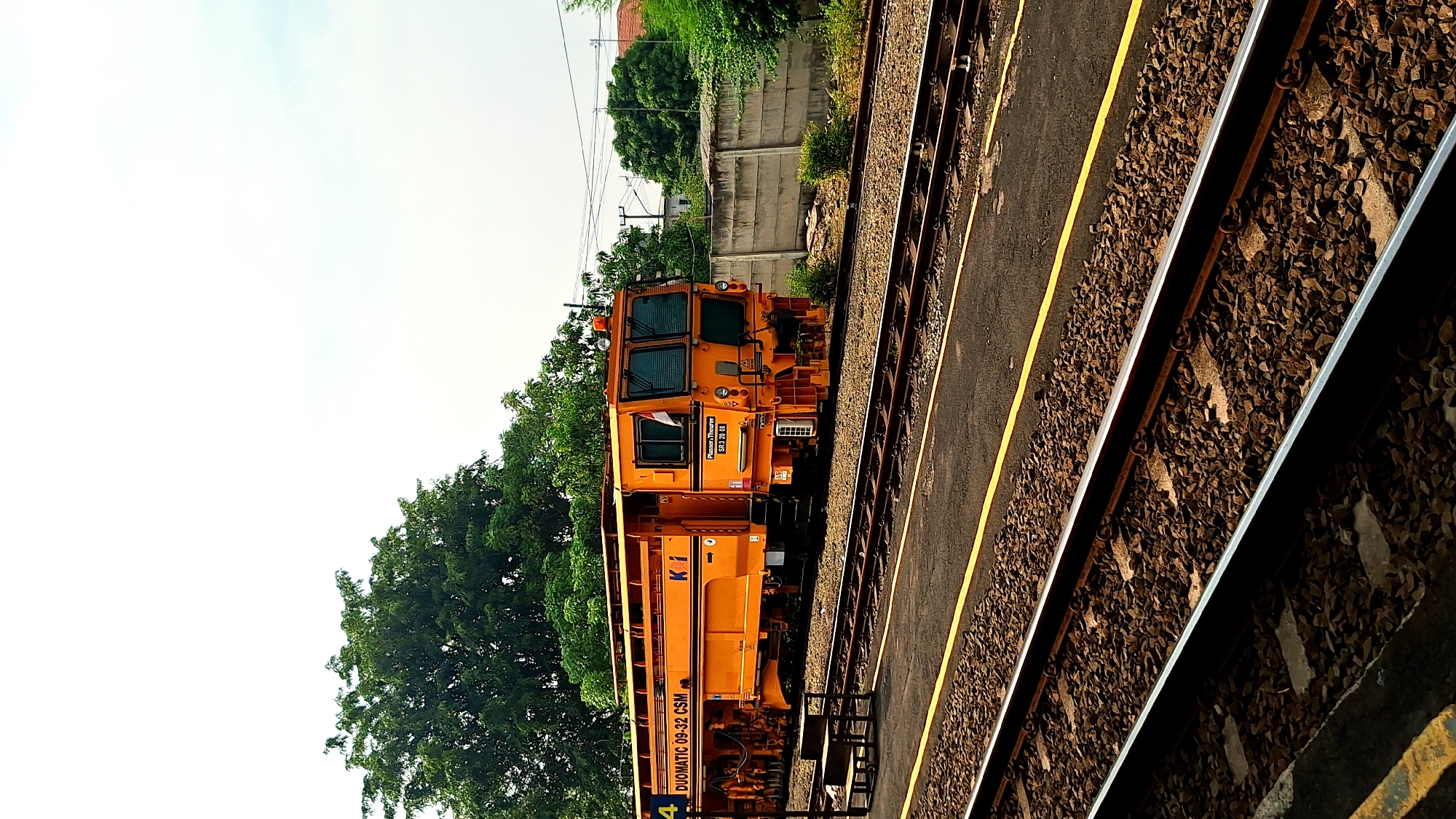 The image shows an orange rail maintenance vehicle, a Plasser & Theurer Duomatic 09-32 CSM, parked on a railway track in Karawang, Indonesia. The vehicle is positioned parallel to the tracks and adjacent to a platform. Beyond the tracks and vehicle, there's greenery, a concrete wall, and a section of sky. The platform edge is visible in the foreground.