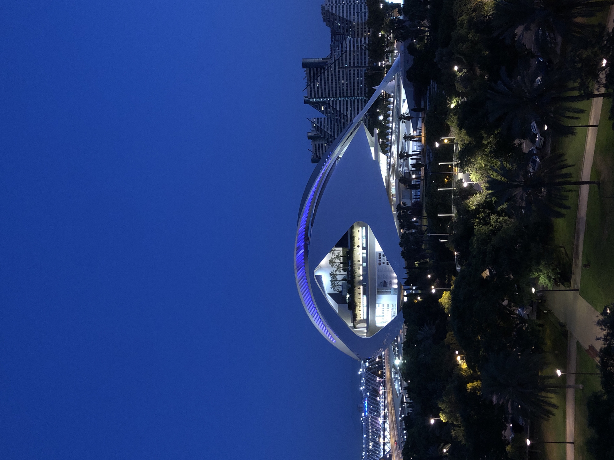 The image captures a twilight view of the City of Arts and Sciences in Valencia, Spain. The striking Hemisfèric building, a contemporary architectural landmark, is prominently featured in the foreground, illuminated with a soft blue light along its upper edges. The building's distinctive eye-shaped structure, designed by Santiago Calatrava, is partially open, revealing internal lighting and a display with the word "ARTS". Palm trees and lush greenery form a dark foreground, with lampposts casting pools of light on pathways. In the background, a cluster of angular, stepped residential buildings with numerous windows are visible, also lit from within. A complex network of illuminated bridges and roads suggests urban infrastructure. The sky is a deep, clear blue, indicative of the fading daylight. There are no people or animals clearly discernible in the scene. The overall impression is one of modern urban landscape at dusk, with a focus on architectural design and lighting.