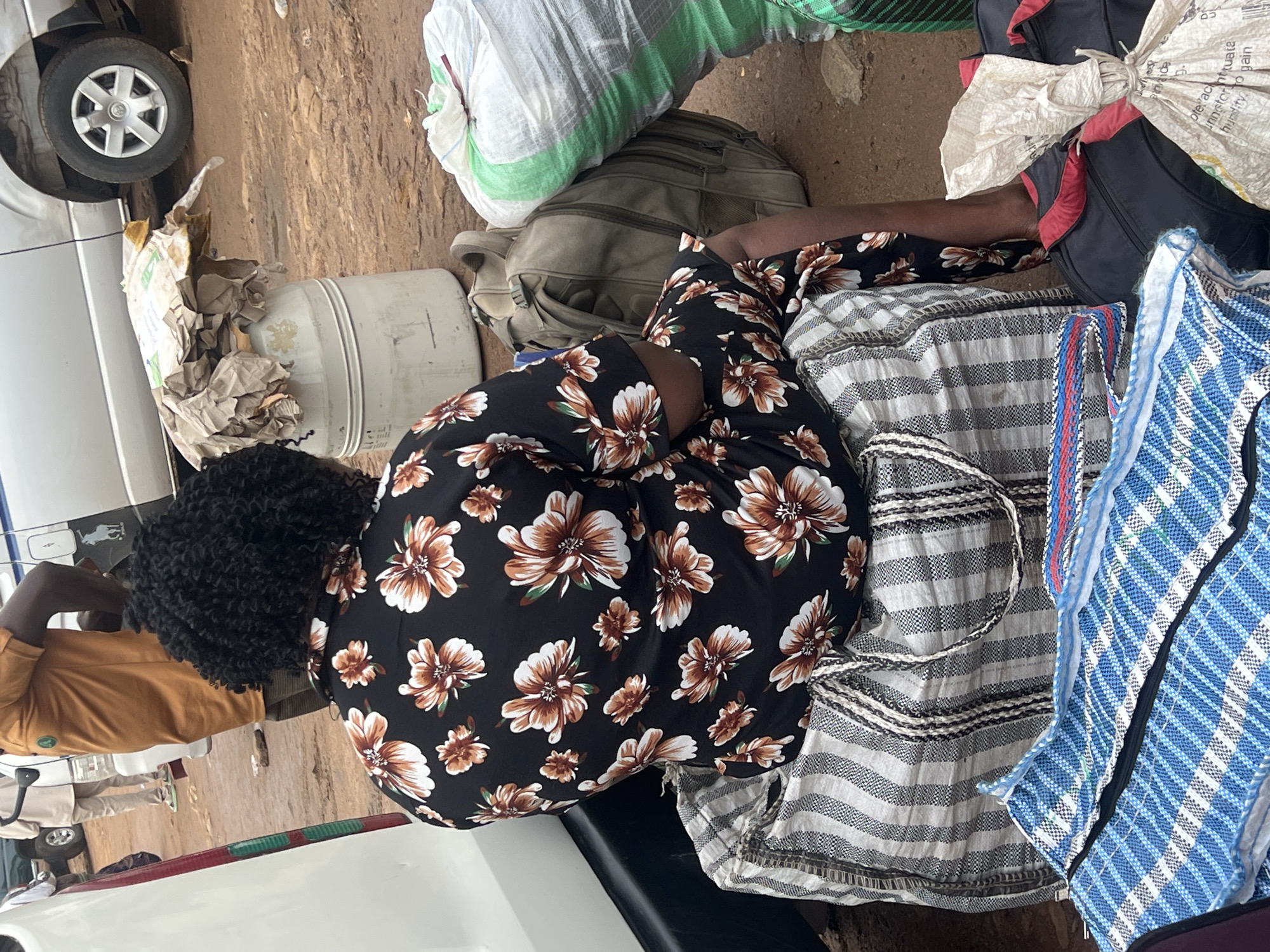 The image shows a woman in a black and floral print dress sitting on the ground next to a white vehicle, surrounded by various bags and objects. She is leaning against a large, dark green backpack. To her left, a light gray, cylindrical object wrapped in brown paper sits near the vehicle's wheel. Several large sacks are visible, including one predominantly white with green stripes, and another smaller, off-white sack tied with string. Adjacent to the woman, on her right, are two large woven bags: one with horizontal grey and white stripes, and another with a blue and white checkered pattern. The ground appears to be dirt or mud, suggesting an outdoor setting, possibly a roadside or market area. The lighting suggests daytime, but the weather is not clearly discernible. The scene appears to be one of transit or preparation for travel, with the woman and her belongings gathered together. No visible text is legible in the image. The overall impression is one of a busy, practical moment, likely in Jos, Nigeria.