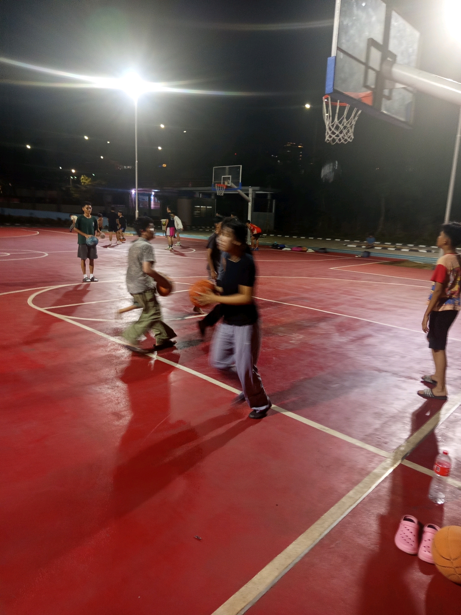 The image captures an outdoor basketball court at night in West Jakarta, Indonesia. A bright overhead light illuminates the red court, casting long shadows from the players. Several individuals are present, engaged in a casual game. One person in the foreground is mid-stride, holding a basketball. To their left, another person stands with a basketball. In the background, other players are visible, along with a basketball hoop. The court appears to be an amenity in a residential area, with trees and buildings visible in the distance. A water bottle and a pair of pink slides are placed on the edge of the court.