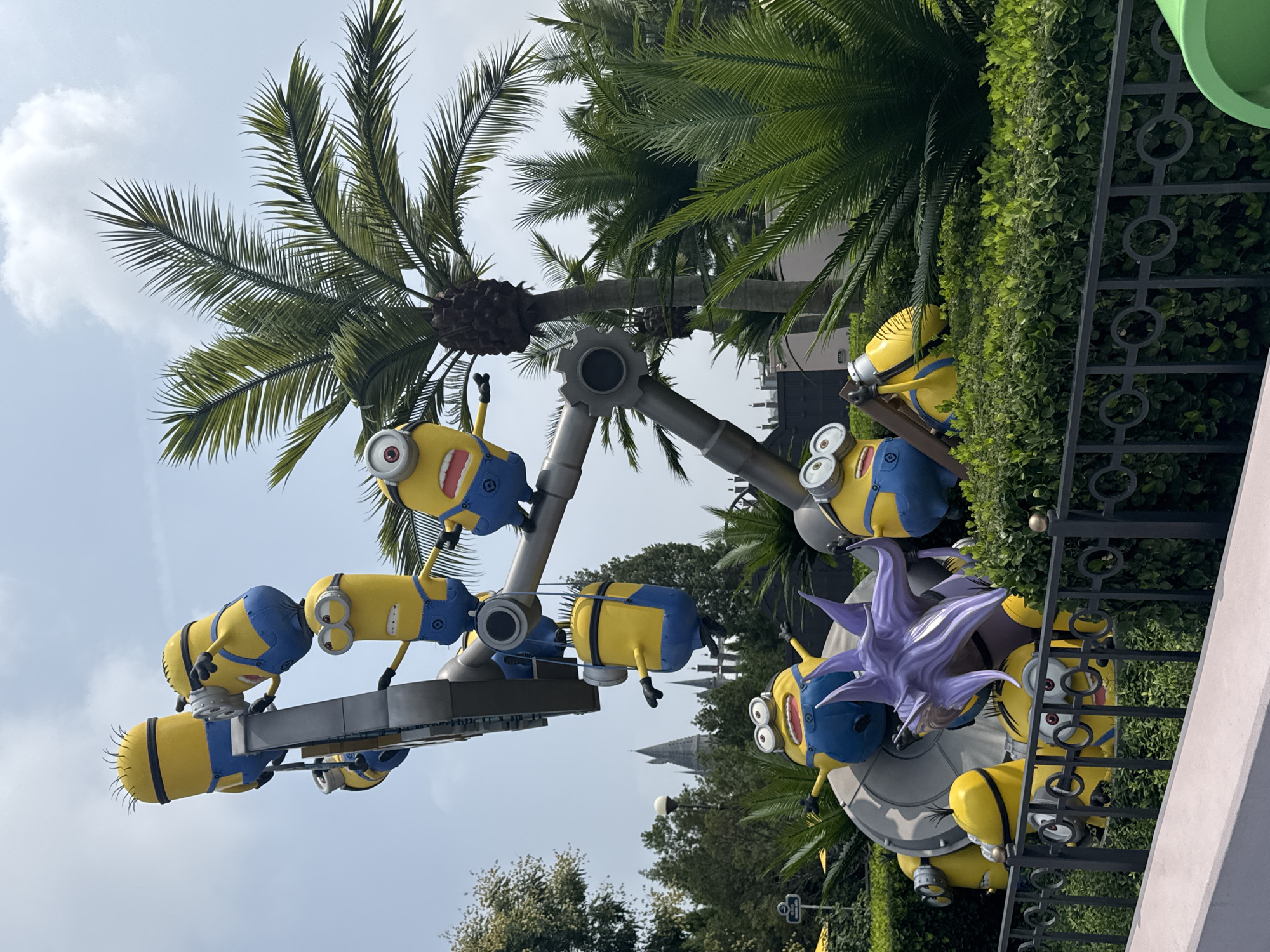 This is a medium shot of a themed amusement park attraction featuring Minion characters from the Despicable Me franchise. The attraction consists of several Minion figures, some appearing to be in motion, mounted on a central metallic structure. In the foreground, a black metal fence separates the viewer from the attraction, behind which is lush green shrubbery. In the background, palm trees and other foliage are visible, along with a hazy sky. A distinctive castle-like structure is visible in the far background, suggesting a larger themed environment. The specific location is identified as Tongzhou District, China.