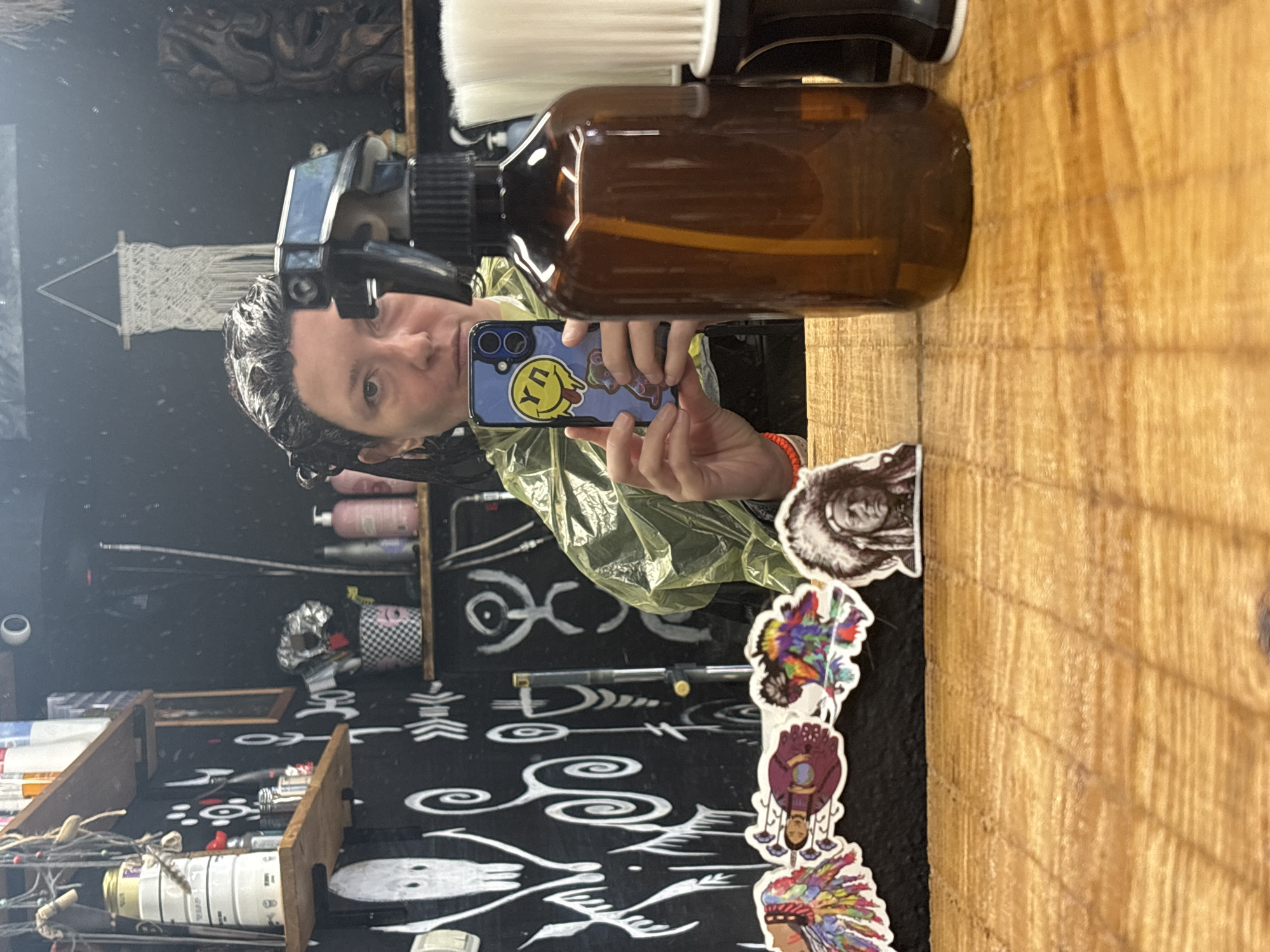 A mirrored selfie in a shop likely located in Bar, Montenegro. The person in the reflection is holding a phone with a sticker-covered case, possibly capturing the image in a mirror. The background is cluttered with shelves stocked with various items and a black and white graphic pattern on the wall. A spray bottle is prominently positioned near the mirror's surface. Several stickers are affixed to the edge of the mirror.