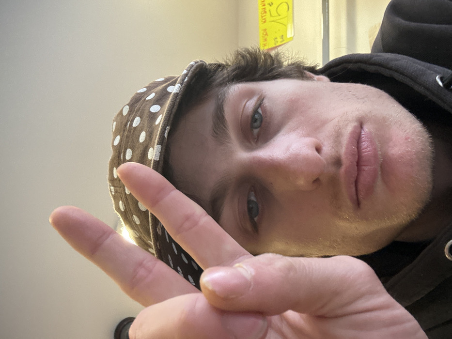 The image depicts a male individual in a close-up, head-and-shoulders orientation. The subject, with light-colored eyes, brown hair, and stubble, wears a dark hooded sweatshirt and a brown baseball cap with white polka dots. His left hand is raised in front of his face, forming a "peace" sign (V-shape with index and middle fingers extended), partially obscuring the left side of his face. The background indicates an indoor environment, with ambient lighting. To the right of the subject's head, a yellow sign is visible against a lighter wall, displaying text including "MAISON KITSUNE" and "15 €". This scene is located in Vincennes, France.