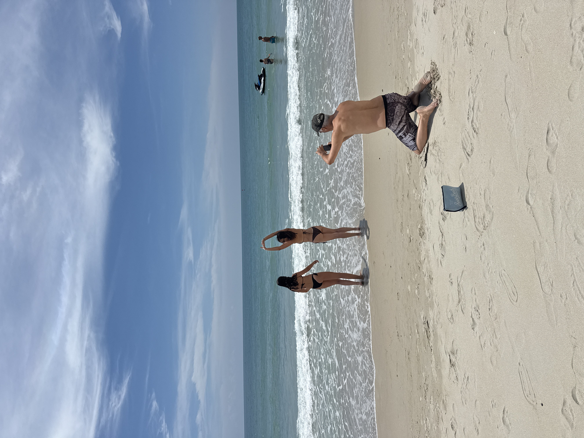 On a sandy beach in the Ko Samui District of Thailand, a man is kneeling on the sand, facing away from the camera, and taking a photograph with his phone. He is wearing a camouflage cap and patterned swim trunks. In front of him, on the sand, is a small blue pouch with the words "St. Barth" printed on it. Two women, both wearing dark bikinis, are standing in the shallow water by the shore, with waves breaking around them. One woman is facing the camera, while the other faces away, with her arms raised above her head. The ocean stretches out to the horizon under a bright blue sky with scattered white clouds. Further out in the ocean, a jet ski is visible, as well as two men standing in the water. The beach is covered in sand with numerous footprints.