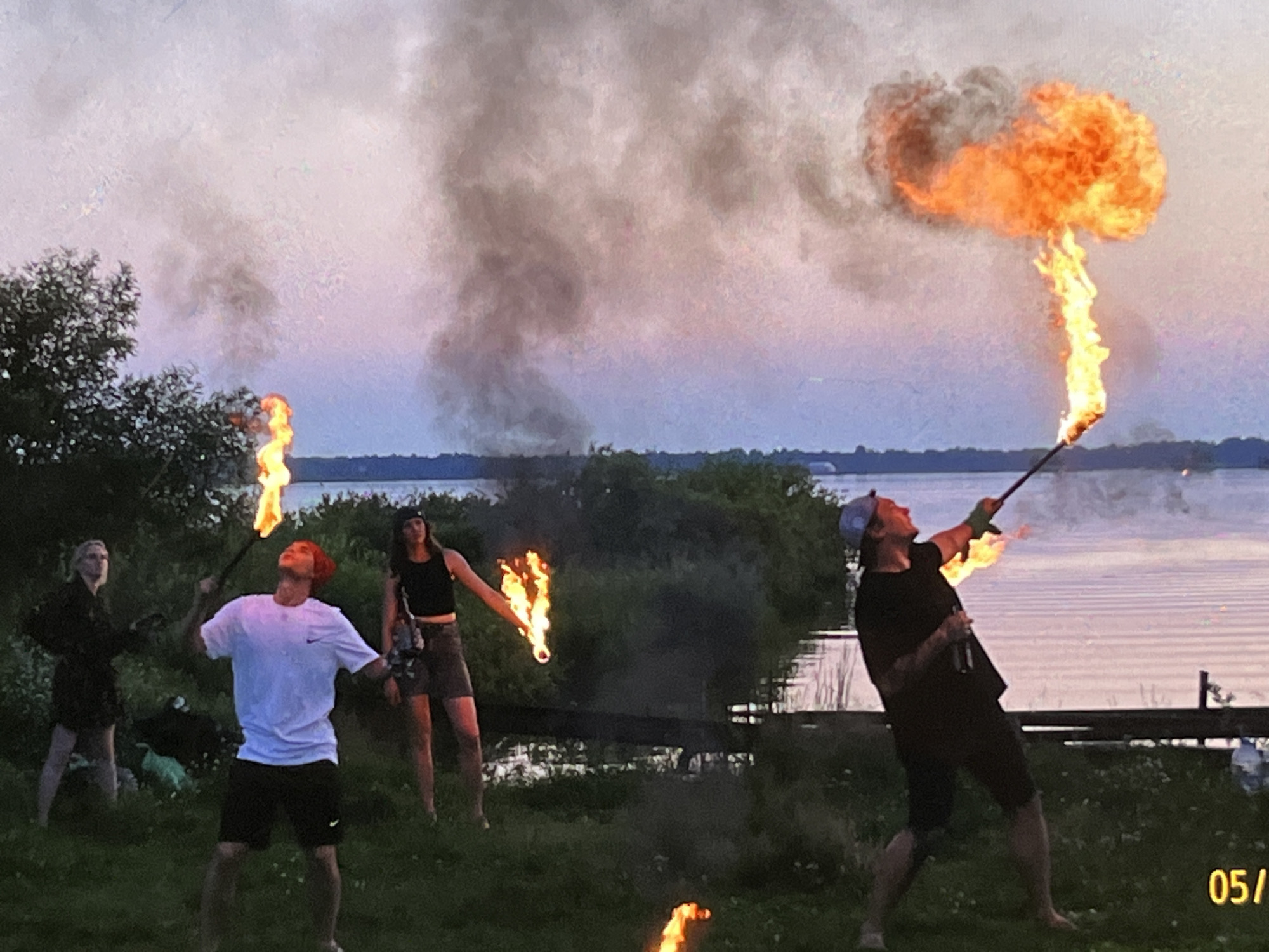 The image depicts a group of people engaging in fire dancing near a body of water, possibly during sunset or twilight. The location is identified as the city of Tyumen, Russia. In the foreground, a man in a white t-shirt and black shorts is holding a flaming object, with flames rising upwards. To his left, a woman in a black top and skirt is also holding a flaming object. Further left, another woman in a black outfit stands looking towards the sky. On the right side of the image, a man in a black t-shirt and shorts is holding a flaming staff, with a large burst of fire and smoke emanating from it. The background features a lake or river with rippling water, and a treeline. Smoke from the fire performance is visible in the sky, tinged with the colors of the setting sun. A date stamp "05/1" is visible in the bottom right corner.