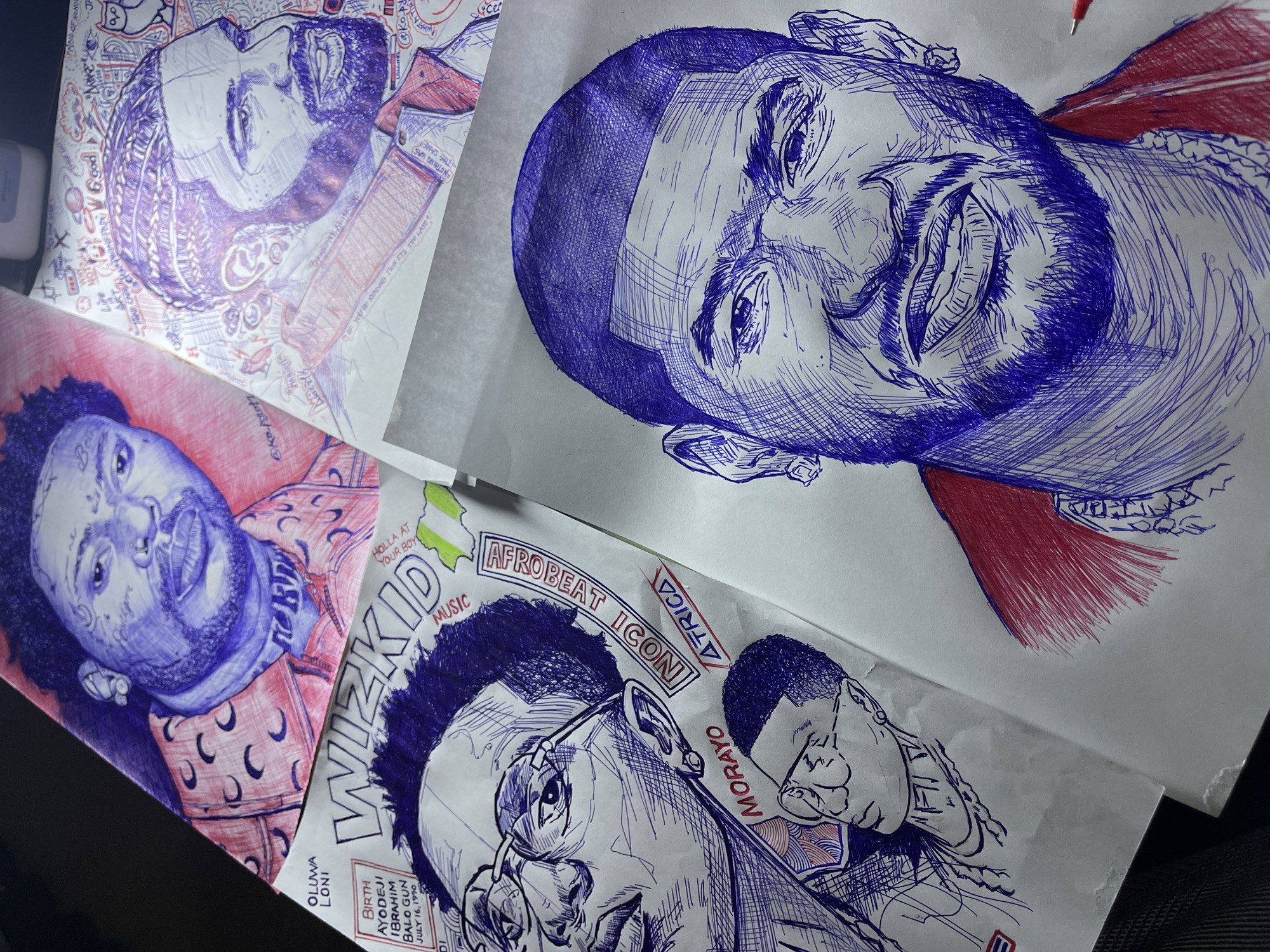 This is a top-down shot of multiple pen-and-ink drawings of various musicians, likely located in Jos, Nigeria. The drawings are on white paper and feature a dominant blue and red color scheme. In the foreground, a detailed portrait of a man with short hair, a beard, and a red jacket is rendered in blue ink. To the left, another drawing shows a man with glasses and braids, accompanied by smaller sketches of other individuals. The background contains a drawing of a man with braids and tattoos, wearing a red patterned shirt, and another drawing of a man with braids and a beard, surrounded by various decorative elements and text. The arrangement suggests these are individual artworks laid out together.