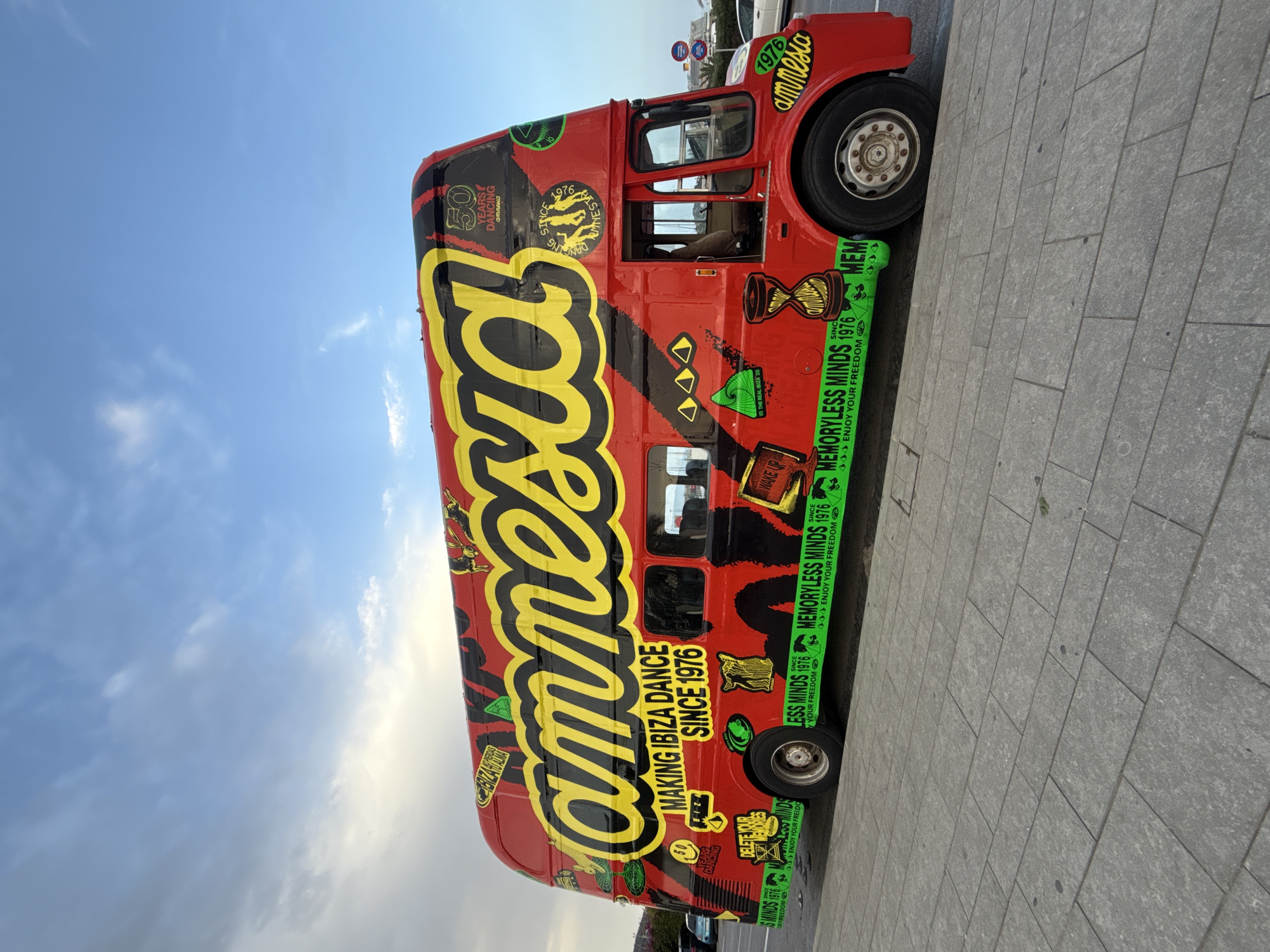 A bright red, double-decker tour bus, heavily decorated with graffiti-style lettering and images, is parked on a sidewalk in Ibiza, Spain. The prominent lettering on the side of the bus reads "amnesia" in large yellow and black font. Below this, text states "MAKING IBIZA DANCE SINCE 1976." The bus also features various graphics, including a smiling face with "50 YEARS DANCING AMNESIA" on the upper deck, an hourglass, geometric shapes, and repeating green text that says "MEMORYLESS MINDS SINCE 1976 ENJOY YOUR FREEDOM." The bus is positioned on a grey, tiled sidewalk next to a street. The background shows a blue sky with scattered clouds and a hint of greenery and distant buildings on the left. The overall scene captures a vibrant, promotional vehicle against an urban backdrop in Ibiza.