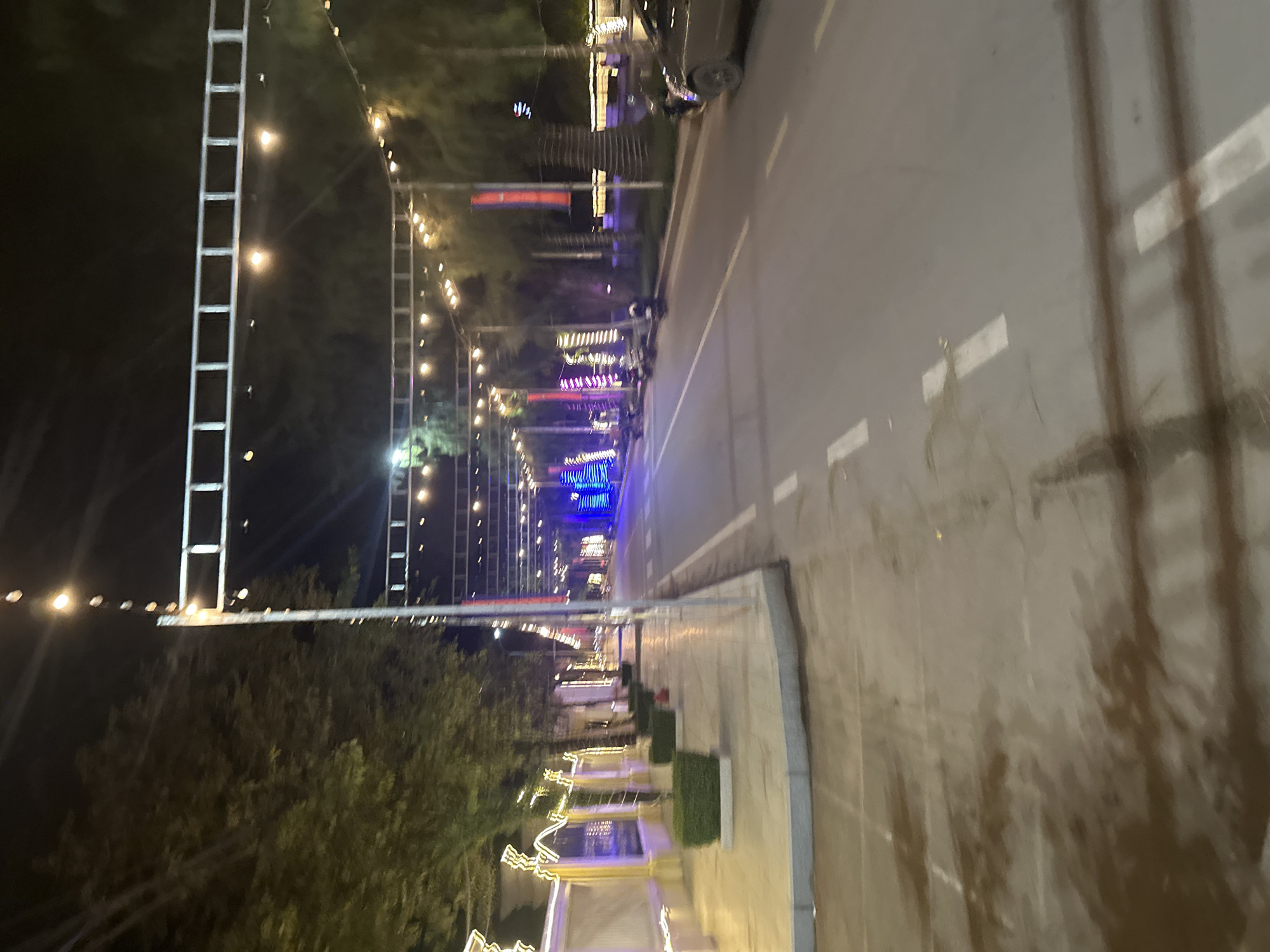 The image depicts a street scene at night in Kampot, Cambodia, likely along the riverside or a main thoroughfare decorated for a festival or holiday. String lights are prominently displayed overhead, suspended by metal structures. The sidewalk on the left is paved and lined with a decorative fence illuminated by yellow lights, with manicured green bushes planted at intervals. Trees are visible along the sidewalk and in the background. The road is paved with visible white lane markings and appears wet, reflecting the ambient light. On the right side of the road, there are parked motorcycles and cars. Further down the street, there are more decorative lights, including blue illuminated structures that resemble Christmas trees, and vertical banners. The overall impression is of a well-decorated urban area at night.