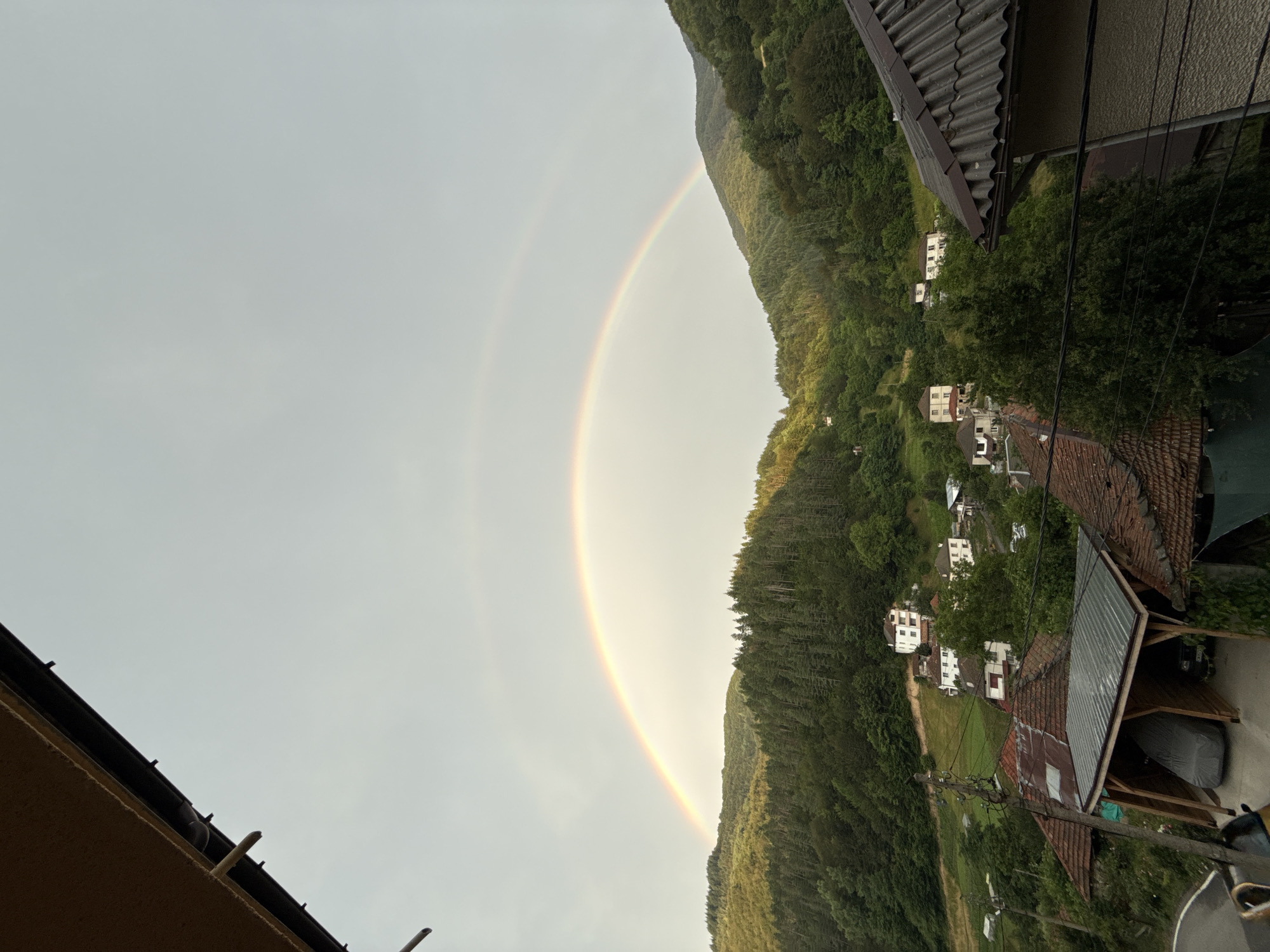 The image depicts a landscape view of Krushev Dol, Bulgaria, featuring a double rainbow arcing across a sky partially obscured by clouds. In the foreground, several rooftops and a covered parking structure with a car are visible. The middle ground showcases a village nestled on a hillside, characterized by scattered houses with red-tiled roofs. The background is dominated by lush, dense green forests covering rolling hills. The lighting suggests it is either early morning or late afternoon, with sunlight illuminating parts of the foliage and the rainbow. No people are present in the frame.