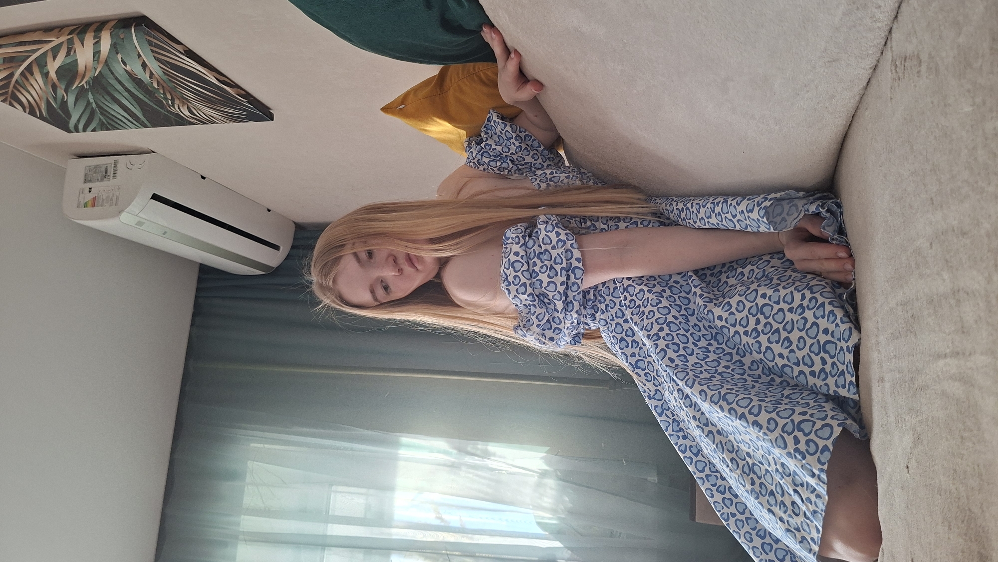 A woman with long blonde hair wearing a blue and white printed dress is lying on a light beige couch, appearing relaxed. She is situated in a room with a green and gold leaf-patterned picture hanging on the wall, next to a white air conditioning unit. In the background, a window with translucent curtains is visible. The location is Chaikovsky, Russia.