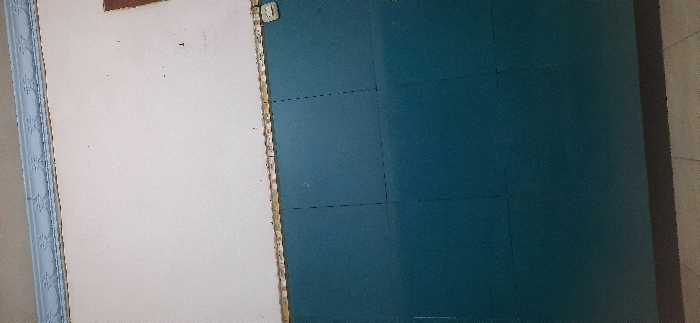 The image shows the interior of a room, possibly a school classroom or office. The walls are painted, the top half is white and the bottom half is teal. There are no people present, and the room is empty except for the walls. There are two thin, black vertical lines dividing the teal wall into three sections. The room has a white ceiling with a decorative, slightly ornate molding. The floor is a light brown tile. There is a small white square object on the right side of the wall near the edge of the image. The coordinates -7.488, 112.715 place the location in the city of Kecamatan Candi, Indonesia.