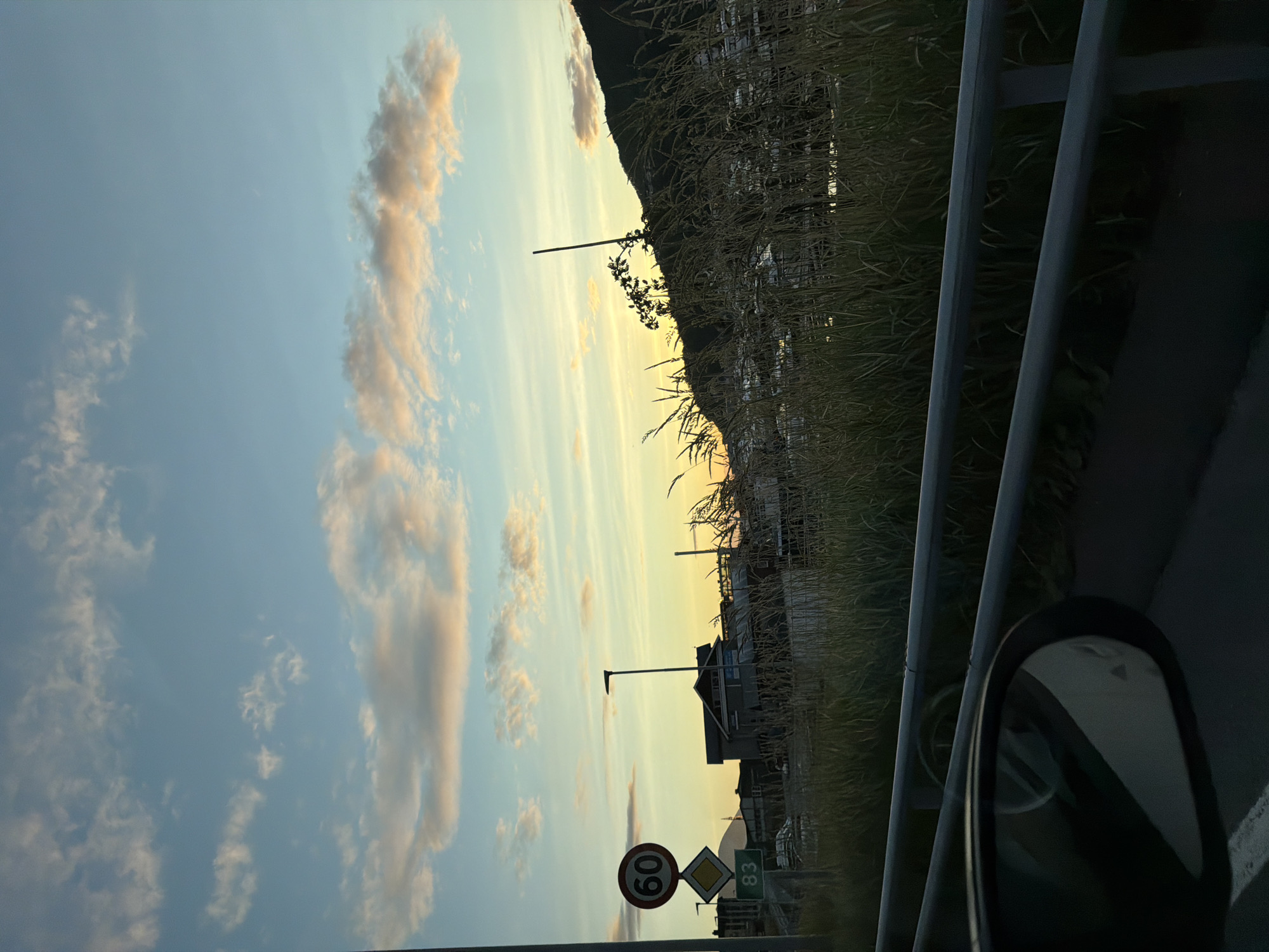 The image depicts a road sign with a speed limit of 60, and another sign below it that is partially obscured, showing the number 83. The scene appears to be a roadside view, likely in Harstad, Norway, given the context. In the background, there are buildings, a street lamp, and a body of water with boats. The sky is a gradient of blue and orange, indicating either sunrise or sunset, with scattered clouds. Tall grass and reeds are visible in the foreground, and a guardrail runs along the side of the road. A car's side mirror is also visible in the lower left corner of the image. The overall impression is of a coastal or harborside area.