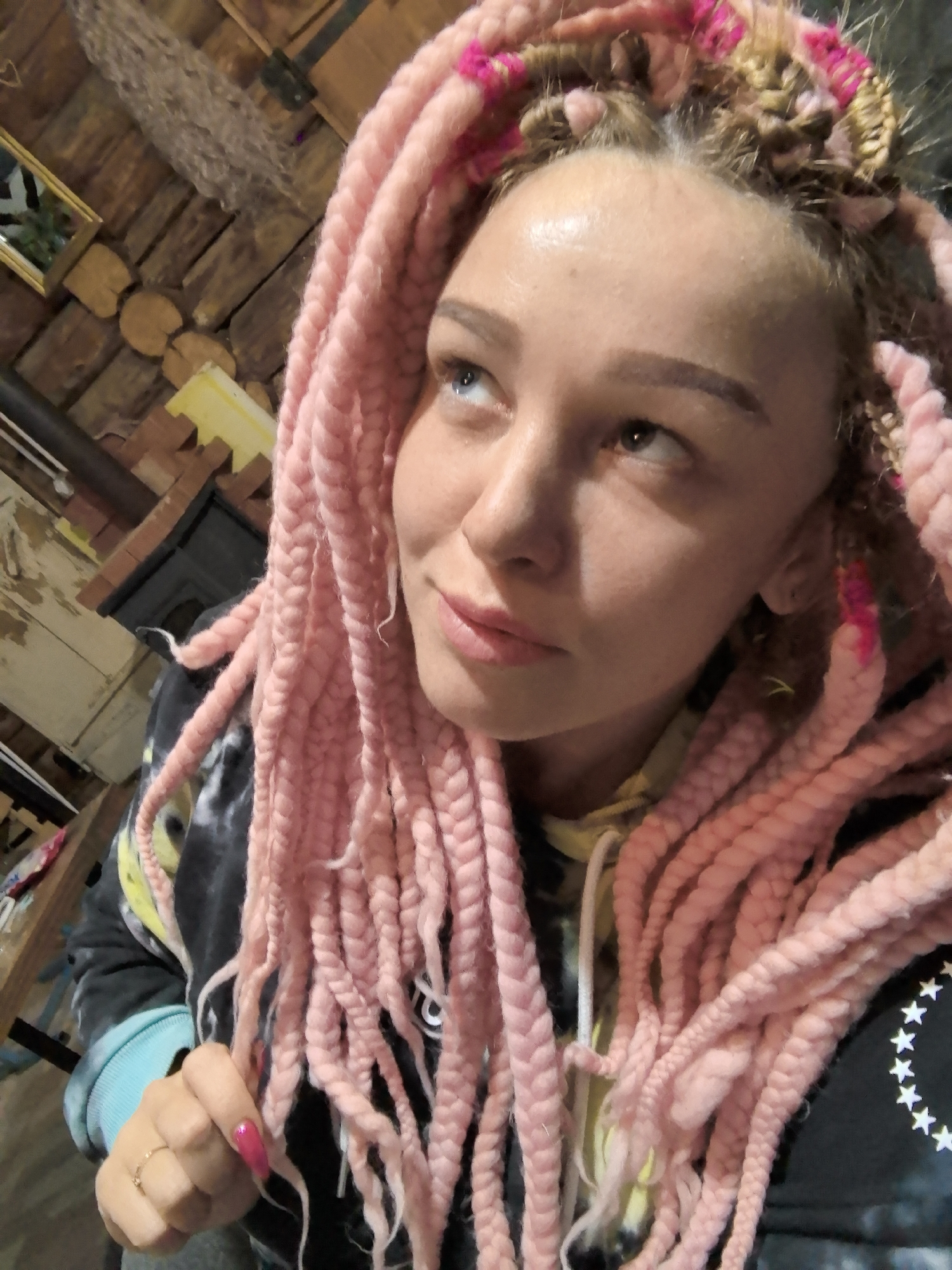 A woman is shown in a close-up shot, looking up and to the left. She has pink braids and fair skin. She is wearing a dark hoodie with a tie-dye pattern and a ring on her finger. The background includes what appears to be a log cabin interior with wooden walls. The image does not contain enough information to verify it was taken in Noviy, Russia.