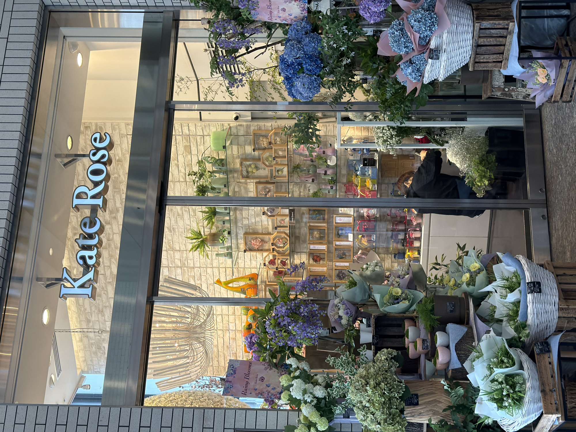 The image shows the storefront of "Kate Rose," a flower shop located in Chiyoda, Japan. The facade features large glass doors and windows, revealing a well-lit interior decorated with a brick-patterned wall. The shop's name, "Kate Rose," is prominently displayed in silver, three-dimensional lettering above the entrance.
Inside, the shop is filled with an abundant display of flowers and plants. Various arrangements are showcased, including large bouquets of blue hydrangeas and purple wisteria-like flowers in the foreground. Baskets filled with white and green flowers are placed on wooden crates, and smaller potted plants and decorative vases are visible on shelves. In the background, framed dried flower arrangements are displayed.
A person, wearing a dark hooded jacket and jeans, is working inside the shop, appearing to arrange flowers. They are positioned behind a counter, with their back to the viewer.
The exterior of the shop is lined with light-colored brickwork, and the entrance has a modern, metallic frame. The overall impression is of a professional and aesthetically pleasing floral business.