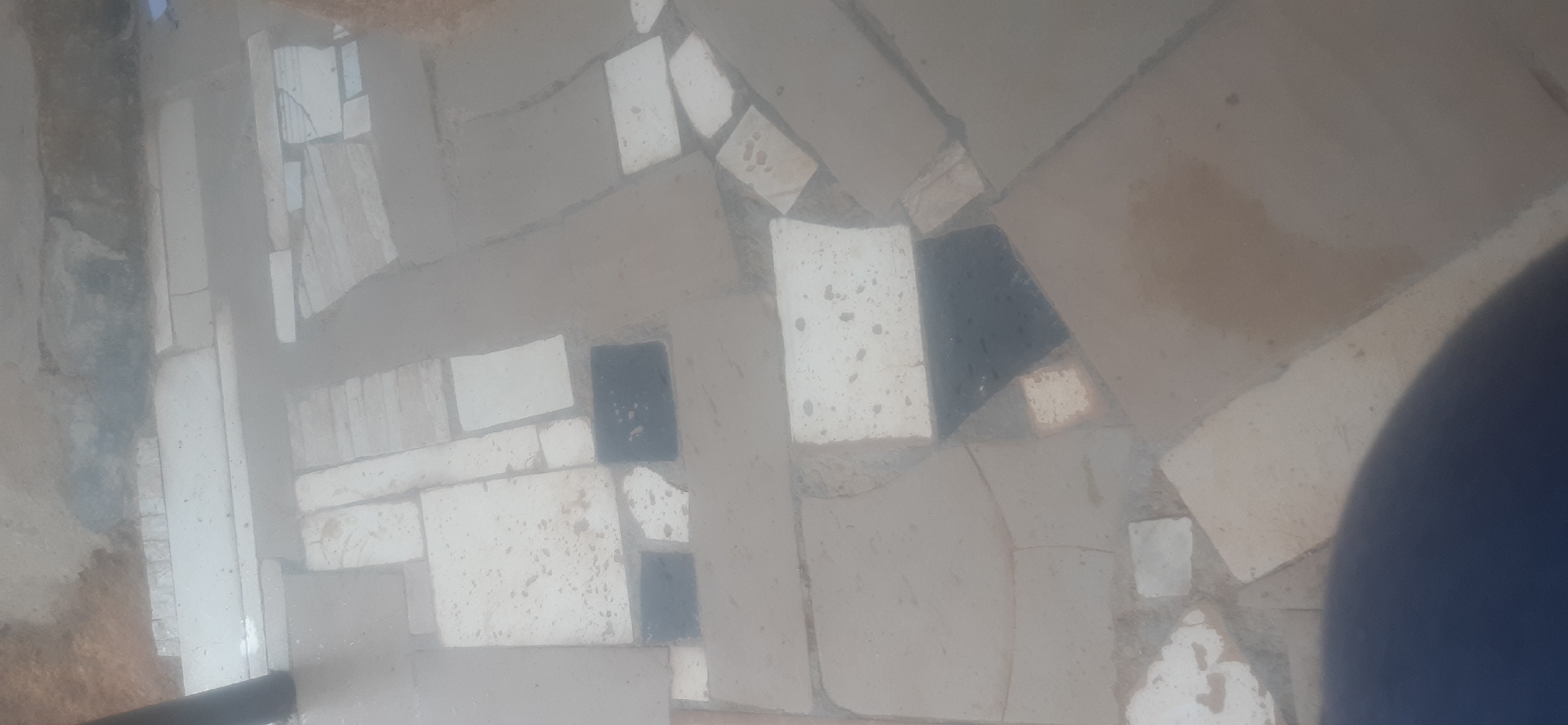 The image shows a paved area with irregular-shaped tiles of varying colors, predominantly off-white and a few dark grey or black tiles. The tiles are laid in a mosaic pattern and appear to be slightly worn and dusty, with some visible grout lines. The lighting is diffuse, suggesting an overcast day or an indoor setting with indirect light. The scene is consistent with a courtyard or walkway in Jos, Nigeria, characterized by its use of locally sourced or common paving materials. The presence of what appears to be a dark, rounded object in the lower left corner might be a shadow or part of an object outside the frame, but it does not detract from the description of the paved area.