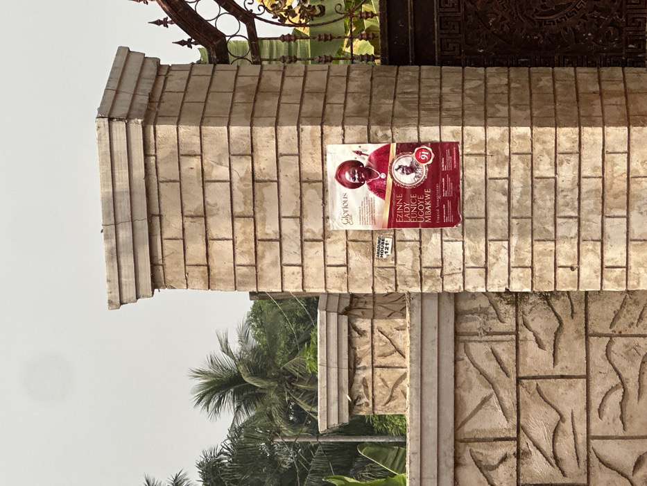 The image displays a light beige, rectangular brick pillar located in Nnobi, Nigeria, serving as a support for an ornate dark metal gate on the right. A reddish-brown "Glorious Exit" funeral announcement poster is affixed to the pillar. The poster features a smiling older woman in a red headwrap and traditional attire, identified as "EZINNE LADY EUNICE UGOYE MBAKWE," above a circular inset image of an older man in a traditional hat, positioned next to a red numeral "91." Below the poster, a white rectangular sign states "AMADUNU HOUSE" and "121." The pillar's cap shows visible cracks. To the left of the main pillar, a lower stone wall with carved patterns extends, beyond which dense green tropical vegetation, including multiple palm trees, is visible against a bright, overcast sky. No individuals or active interactions are visible in the scene.