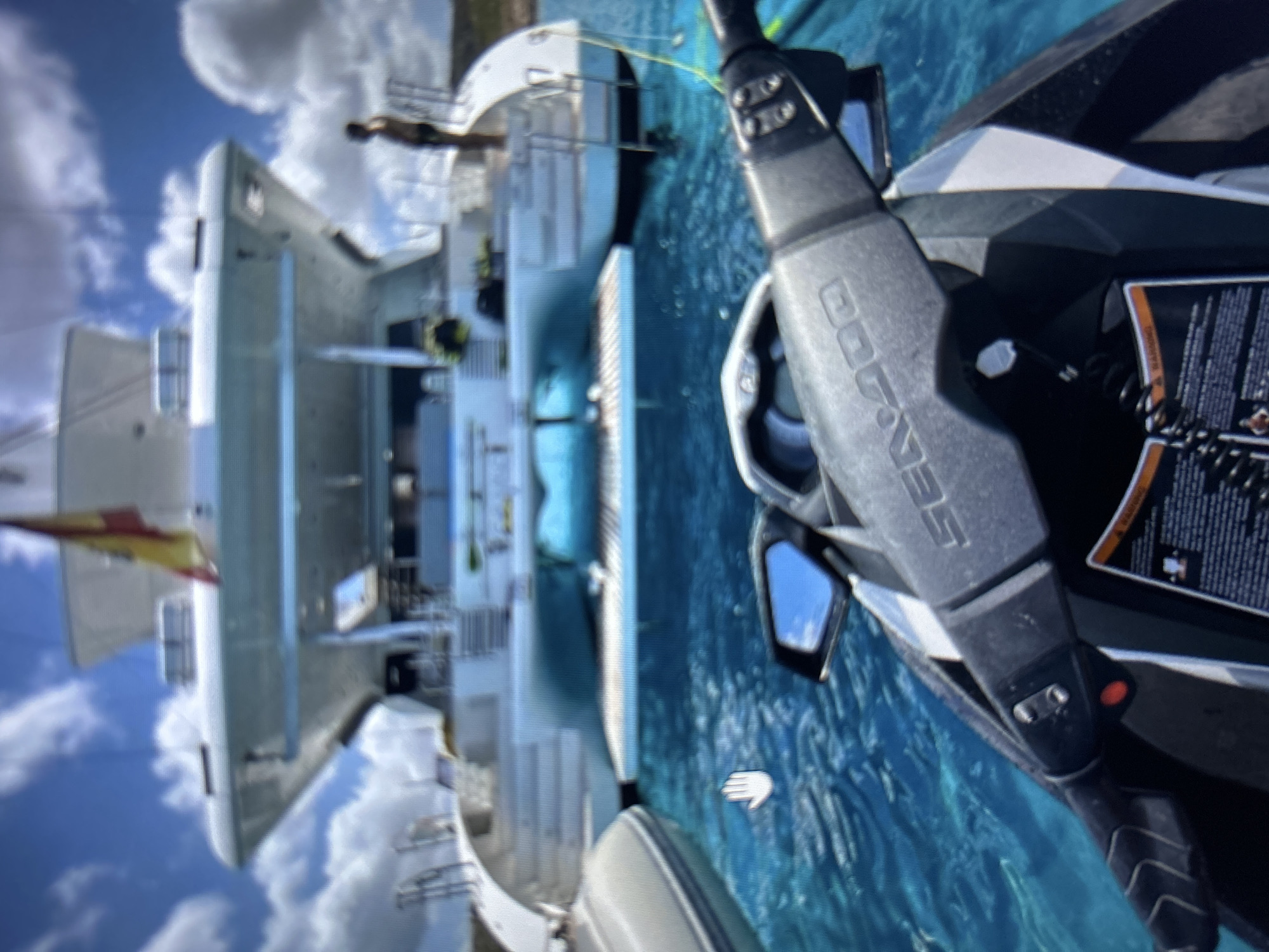 A low-angle perspective from the front of a personal watercraft (Sea-Doo jet ski) captures the stern of a large, white catamaran yacht anchored in clear, turquoise water under a partly cloudy sky. The jet ski's handlebars and part of its body are in the foreground, with a partially visible warning label and a side-mirror reflecting sky. A white hand cursor overlay is present in the water to the left of the jet ski.
In the midground, the catamaran's wide stern is prominent, featuring a lowered swim platform directly above the water, equipped with ladders and railings for water access. The yacht's submerged hulls appear light blue. A grey inflatable dinghy is partially visible floating to the left of the catamaran. A person is standing on the upper stern deck of the catamaran, facing away from the camera, wearing dark swim trunks. An inflatable object, possibly a water toy or fender, is visible between the catamaran's hulls at the stern. A red and yellow flag, identifiable as the flag of Spain, flies from a flagpole on the yacht's starboard side. The background consists of a bright blue sky with scattered white clouds, and a faint landmass is visible on the horizon to the right. The flag on the yacht indicates a connection to Spain. No visual information within the image confirms the specific geographic location as the city of Burgas, Bulgaria.