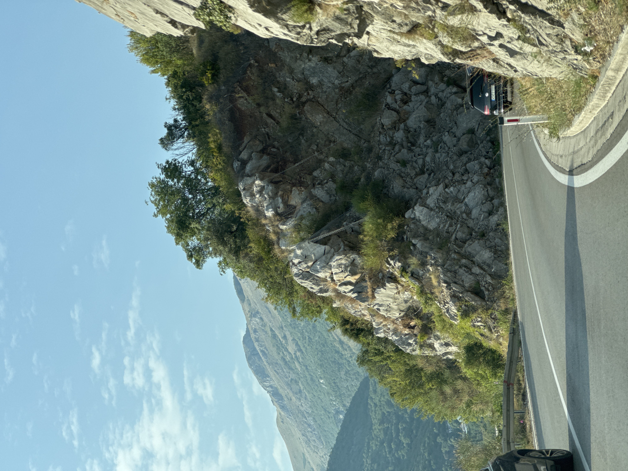 The image depicts a scenic mountain road in Košljun, Montenegro. A black Volkswagen Golf is parked on the right side of the road, partially obscured by a rocky cliff face. The road curves to the right, with a white line marking the lane separation and a curb on the right edge. Vegetation, including trees and bushes, grows along the rocky slopes of the mountain. In the background, rolling green hills and mountains stretch towards a clear blue sky with scattered white clouds. The overall scene is one of natural beauty and tranquility, with the road appearing to be a well-traveled route through the mountainous terrain.