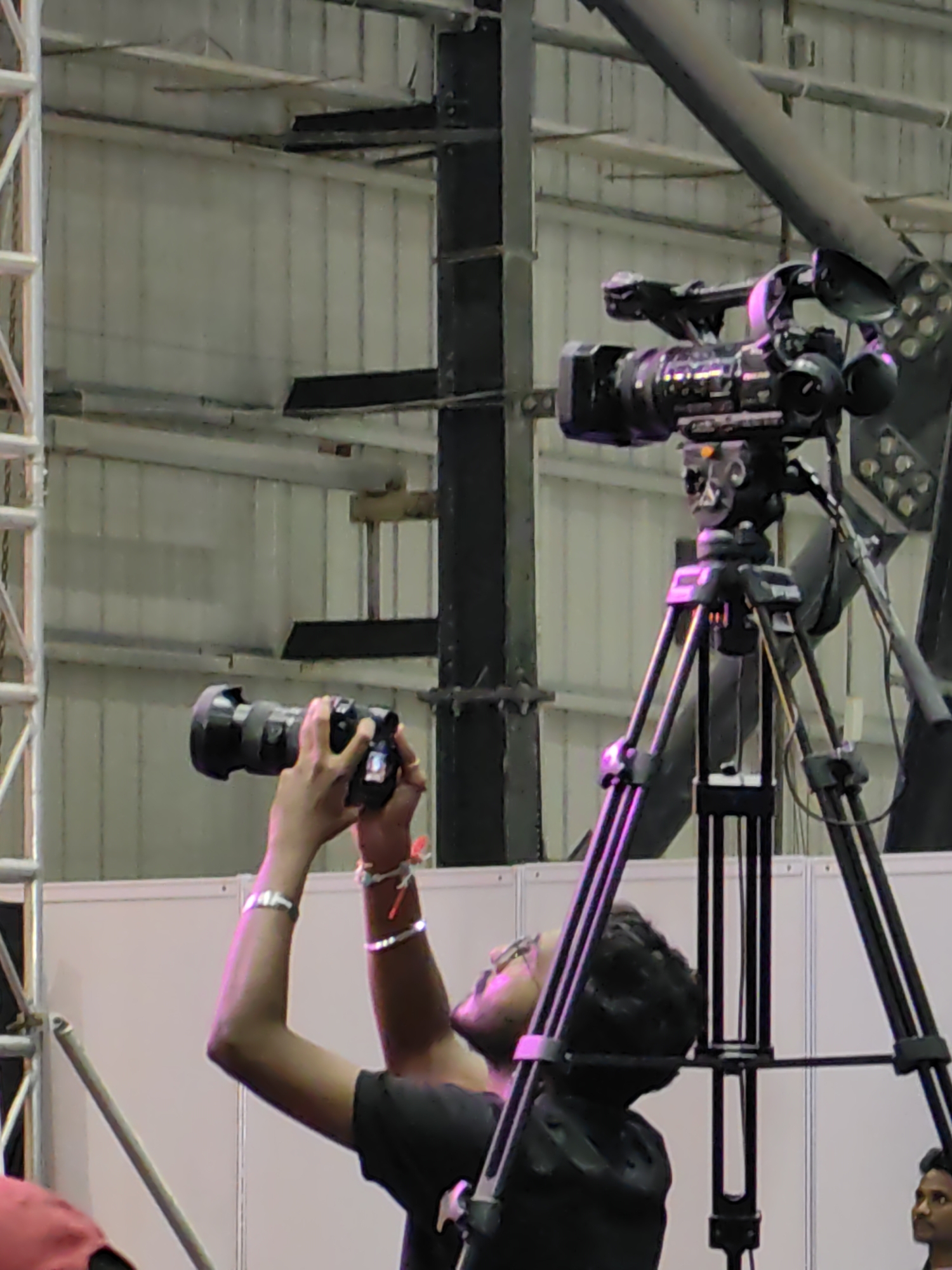 In Mumbai, India, a person is using a handheld camera. Another person is operating a professional video camera mounted on a tripod. The scene appears to be indoors, possibly at an event or venue, with industrial-style metal structures and lighting visible in the background. The overall lighting is somewhat dim, with some pinkish hues from stage lights.