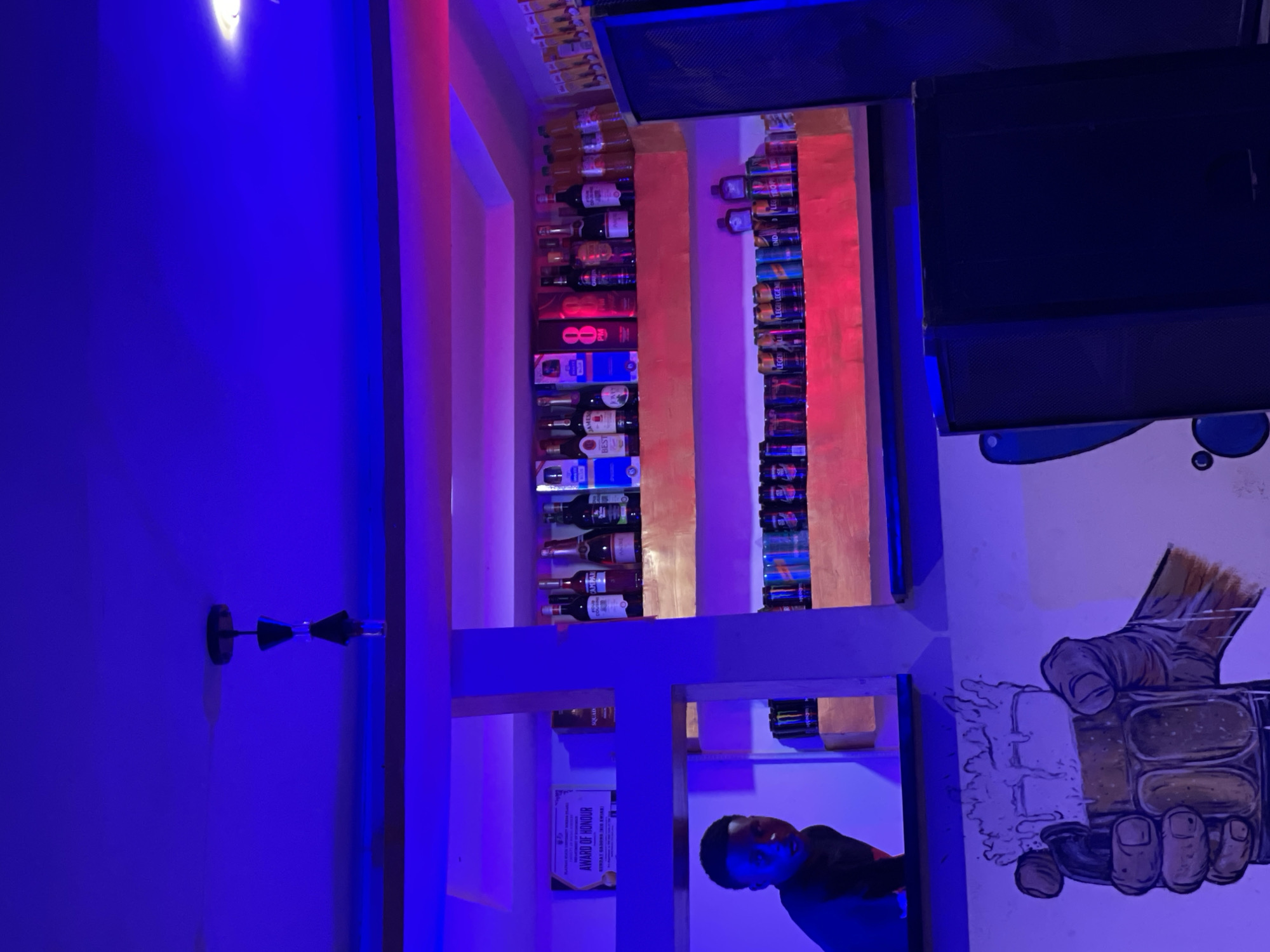 A man is present in a dimly lit room with blue and pink lighting. The room appears to be a bar or a lounge, with shelves displaying various bottles of alcohol. The shelves are made of wood and are illuminated by colored lights, creating a vibrant atmosphere. In the background, there is a mural on the wall depicting a hand holding a frothy drink. The man, who is partially visible in the frame, seems to be looking away from the camera. There is text visible on a certificate or award hanging on the wall, which reads "AWARD OF HONOUR" and "SQUAD". Other visible text includes brand names such as "CAMPARI", "JAMESON", and "PM". The overall impression is that of a lively, modern establishment. The time of day is likely evening or night due to the artificial lighting.