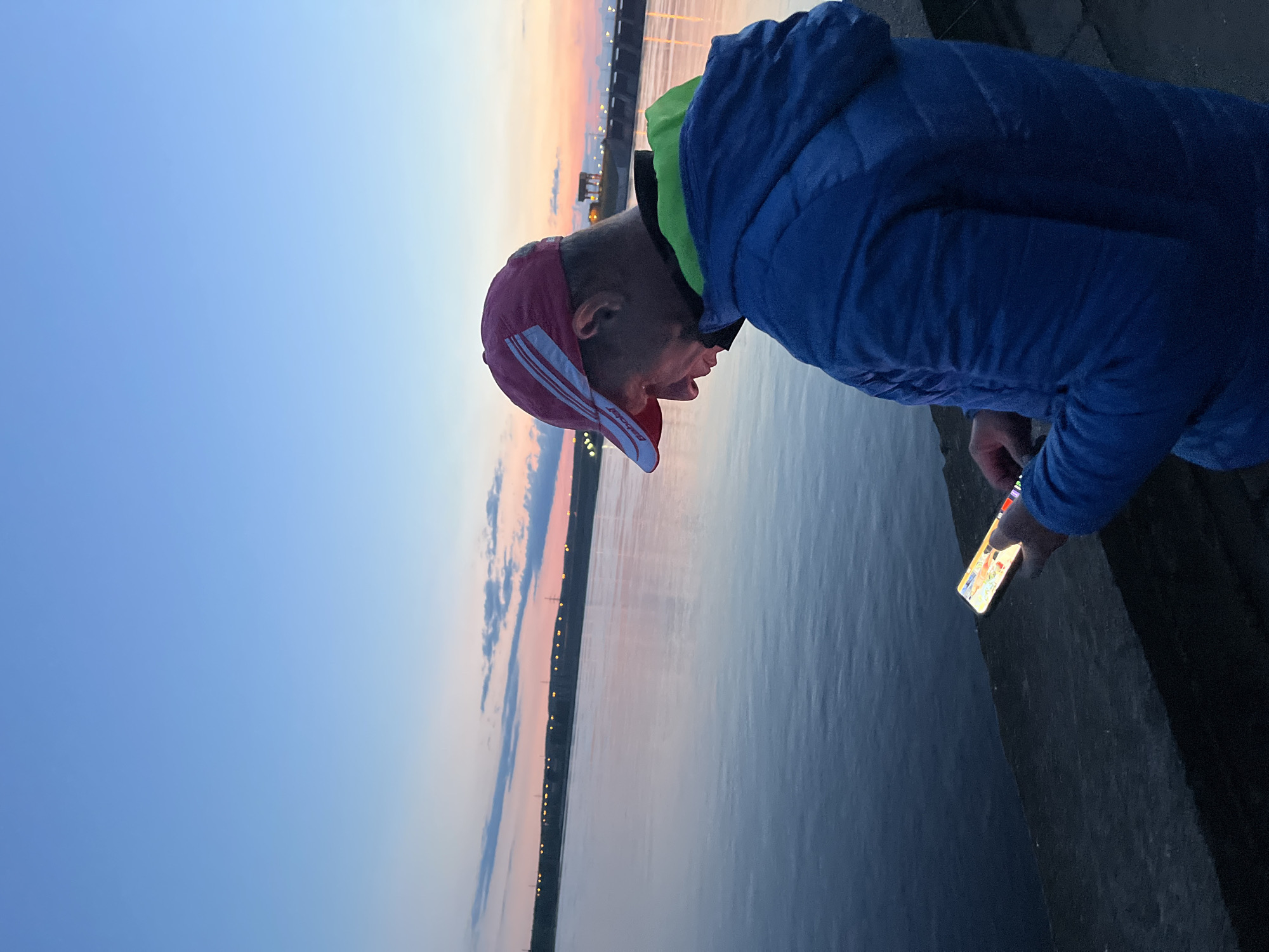 The image captures a man standing on an embankment beside a wide body of water during twilight. The sky transitions from a light blue overhead to warm pink and orange hues at the horizon, indicative of either sunrise or sunset. Distant artificial lights punctuate the far shoreline, suggesting the presence of a town or structures. The water's surface appears calm, reflecting the ambient light.
The man, shown from the side, is leaning forward and looking intently downwards at a brightly illuminated smartphone held in his right hand. He is wearing a blue hooded jacket over a visible green layer and a red and white baseball cap. The brand "BABOLAT" is clearly printed on the white stripe of his cap. His mouth is slightly open, suggesting concentration or a momentary reaction to what he sees on the phone. The phone's screen casts a noticeable glow onto the water immediately below where he stands. The setting aligns with Chaikovsky, Russia, on a major waterway.
The primary activity involves the man's focused interaction with his smartphone against the picturesque backdrop of the serene water and twilight sky. His posture suggests deep engagement with the device, possibly observing something in the water reflected by the phone's light or simply absorbed in its content. The quiet atmosphere and the soft light create a peaceful, contemplative mood.