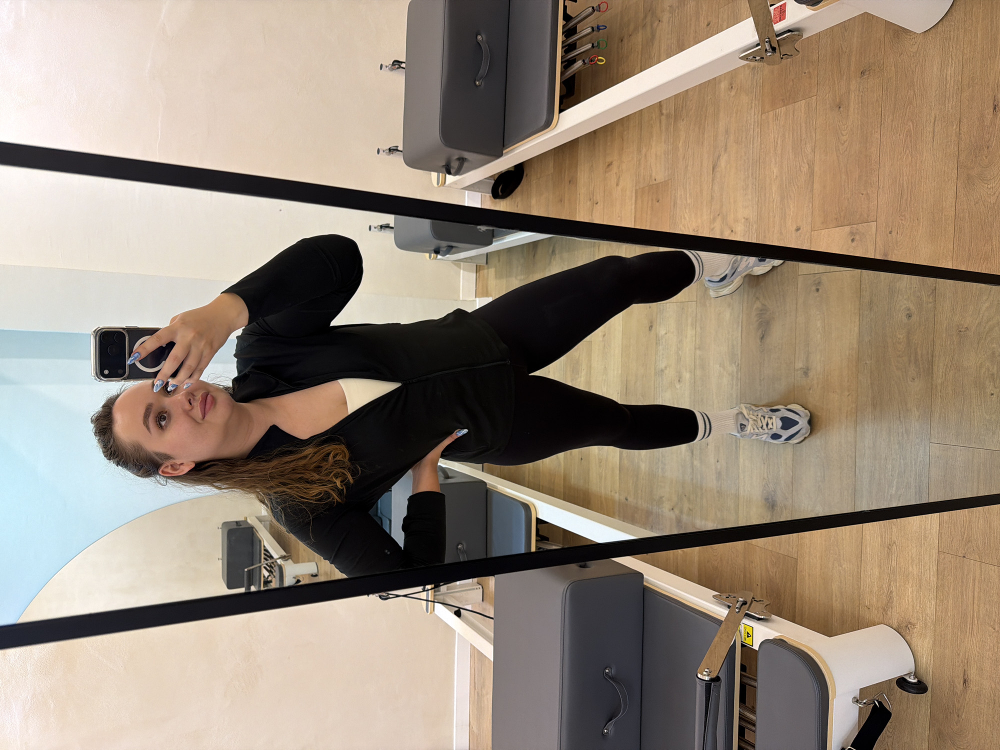 A young woman is captured in a mirror selfie, standing sideways in what appears to be a pilates studio. She holds a smartphone with a clear case and a circular black ring holder in her right hand, positioned to take the photo. Her long, brown hair is mostly pulled back, and she looks directly towards her phone's screen with a neutral to slightly smiling expression.
She is dressed in activewear, consisting of a black long-sleeved top worn over a white sports bra or top, black leggings, and white athletic shoes with blue and gray accents, paired with white socks that have black stripes. Her fingernails are painted with a blue and white design.
The setting is an indoor space with light wooden flooring. Two pilates reformer machines are visible in the reflection, one prominent in the foreground and another partially seen further back. Both reformers have gray padded surfaces and white frames. The reformer in the foreground shows springs and part of its mechanism. The walls in the reflection are a light off-white color, with one section transitioning to a soft light blue or aqua color with a curved boundary.
The overall atmosphere suggests a bright, clean, and modern fitness environment. No specific time of day or weather is discernible, but the indoor lighting is even. The activity is clearly a mirror selfie, posing in a fitness studio in Ramat Gan, Israel. No large, readable text is visible on the equipment or walls.
