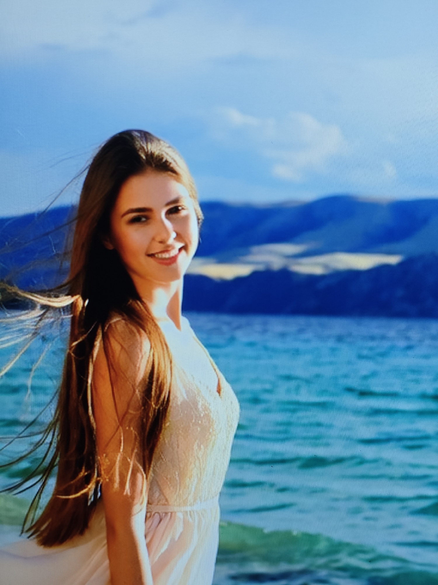 A young woman with long, brown hair flowing in the wind stands on the shore of a body of water, likely a lake or ocean. She is wearing a light-colored, possibly cream-colored, dress with a v-neckline. Her hair is illuminated by sunlight, creating a warm glow. The background features a vast expanse of blue water with waves and distant mountains under a bright, partly cloudy blue sky. The woman is smiling and looking towards the viewer, positioned on the left side of the frame with her body angled to the right. The overall impression is one of natural beauty and serene outdoor activity.
